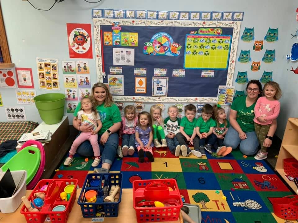 Two adults and several children sitting on a colorful rug in a classroom