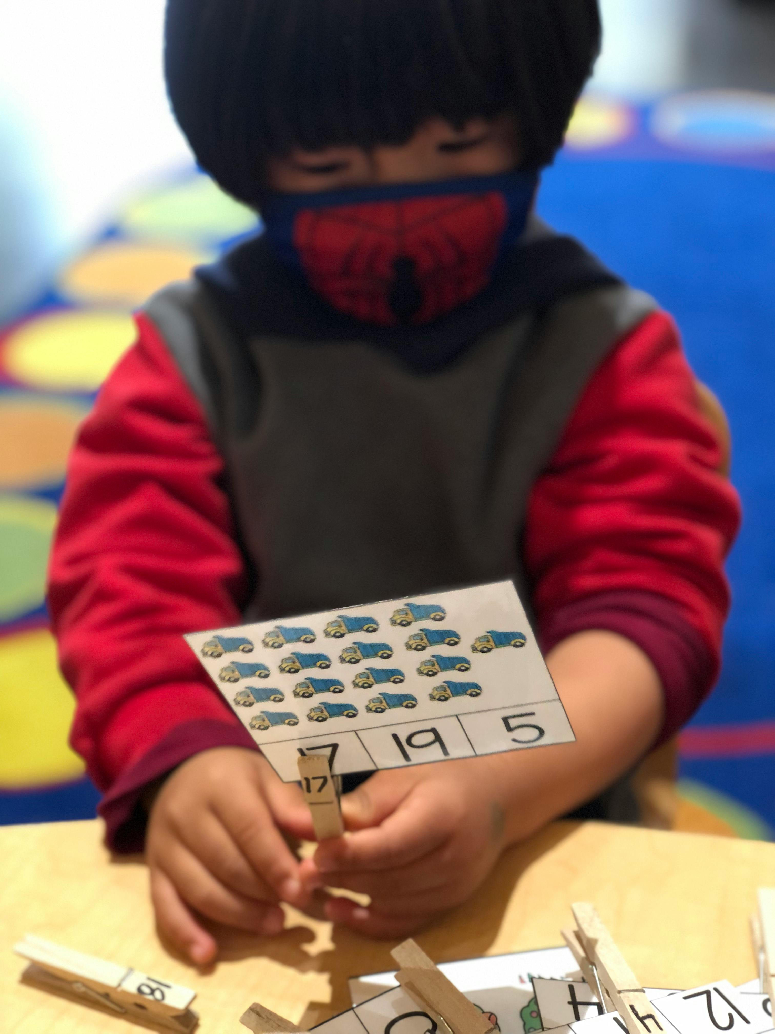 Child in a mask holding a card with car images and numbers, using a clothespin
