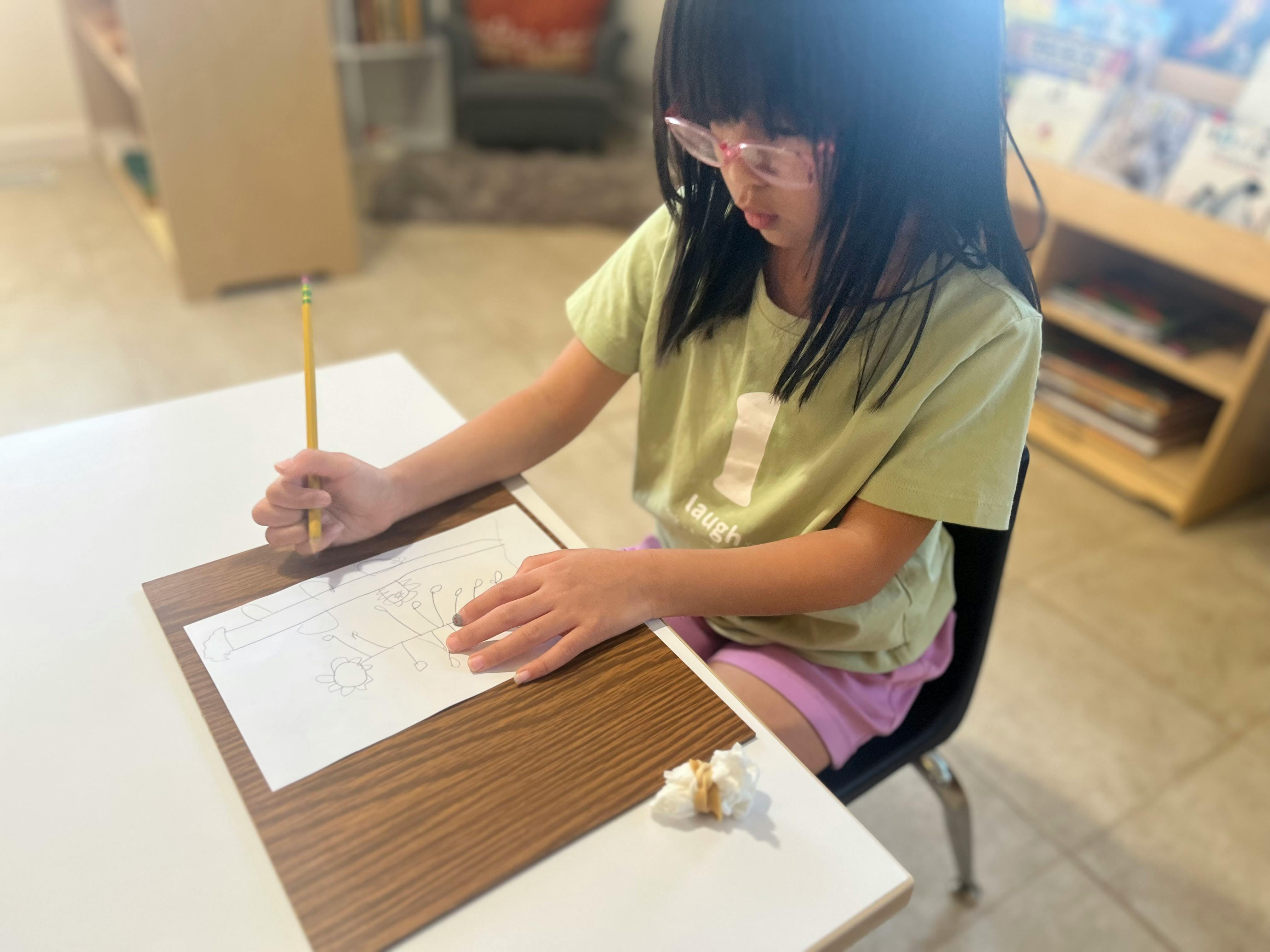 Child with glasses drawing on paper with a pencil at a desk
