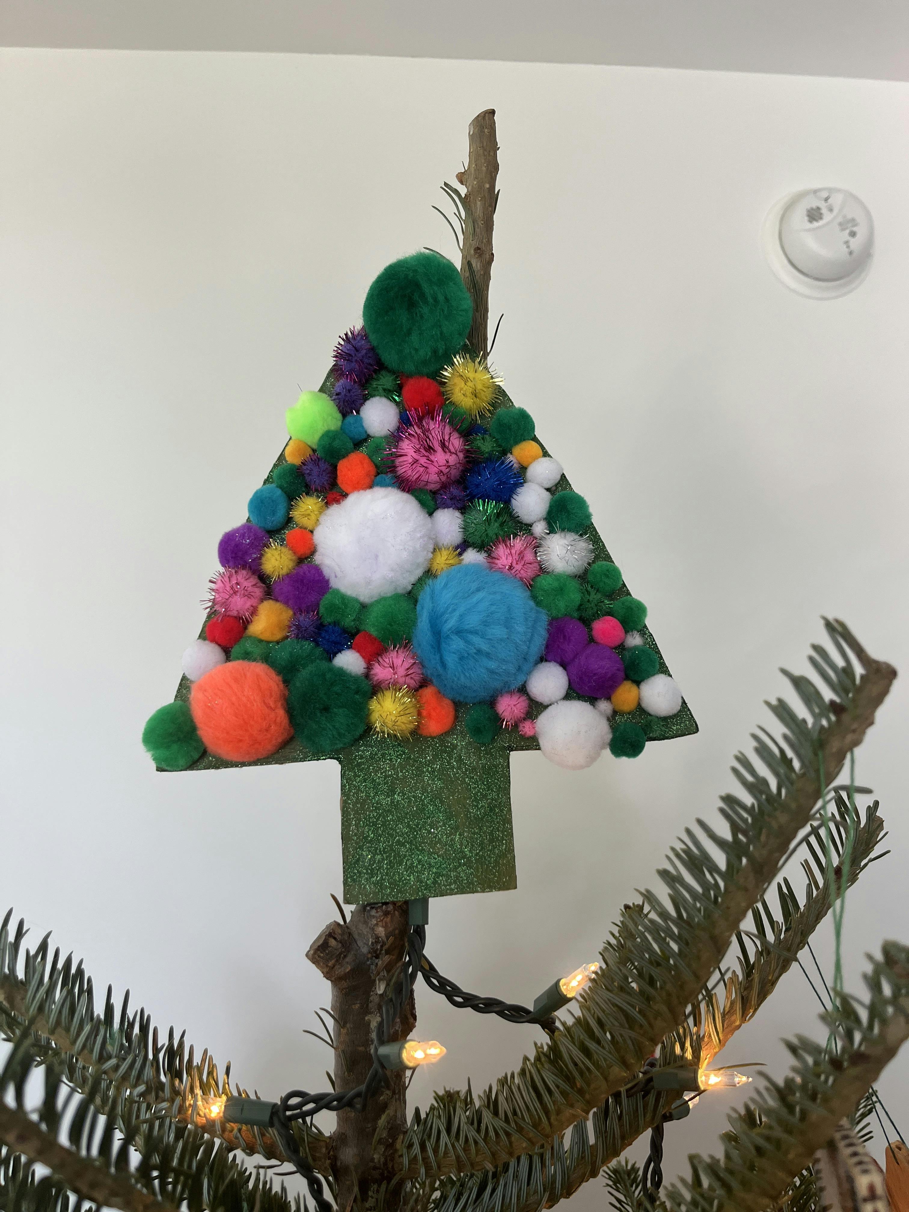 Decorative triangular tree topper covered in colorful pom poms on a tree