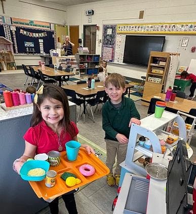 Child holding a tray of play food with another child standing beside a toy kitchen