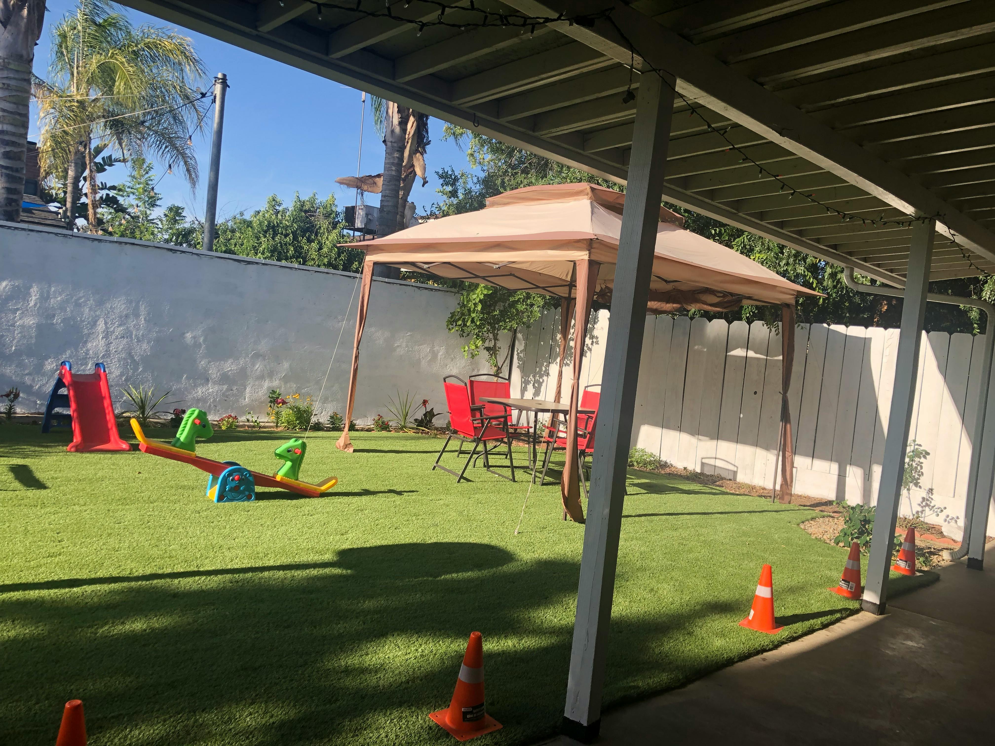 Backyard with artificial grass, a red slide, a seesaw, and a gazebo with red chairs