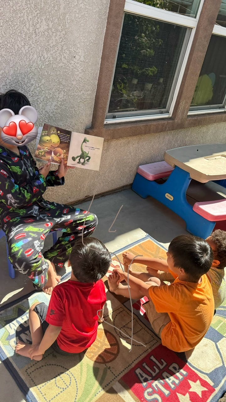 Person reading a book to three children sitting on a rug outdoors