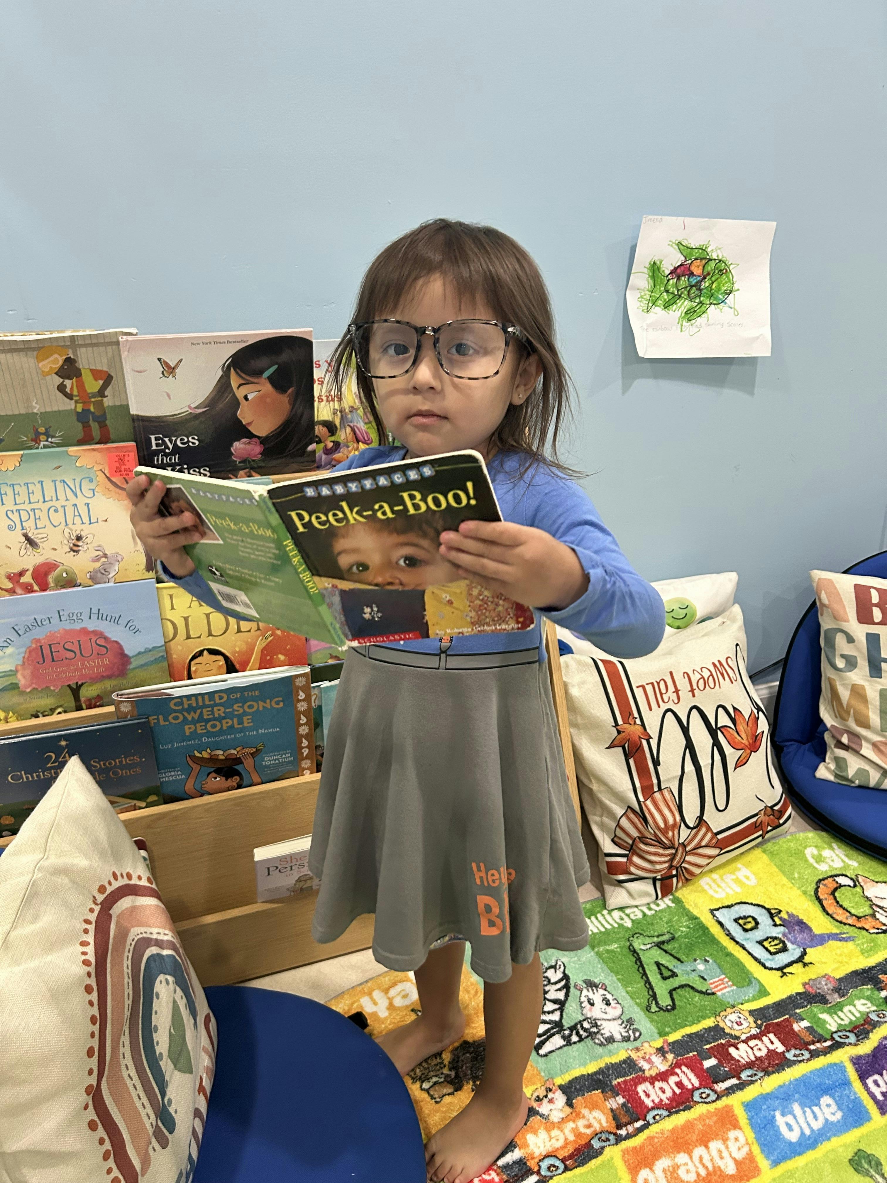 Child wearing glasses and a gray skirt, holding a book titled "Peek-a-Boo!"