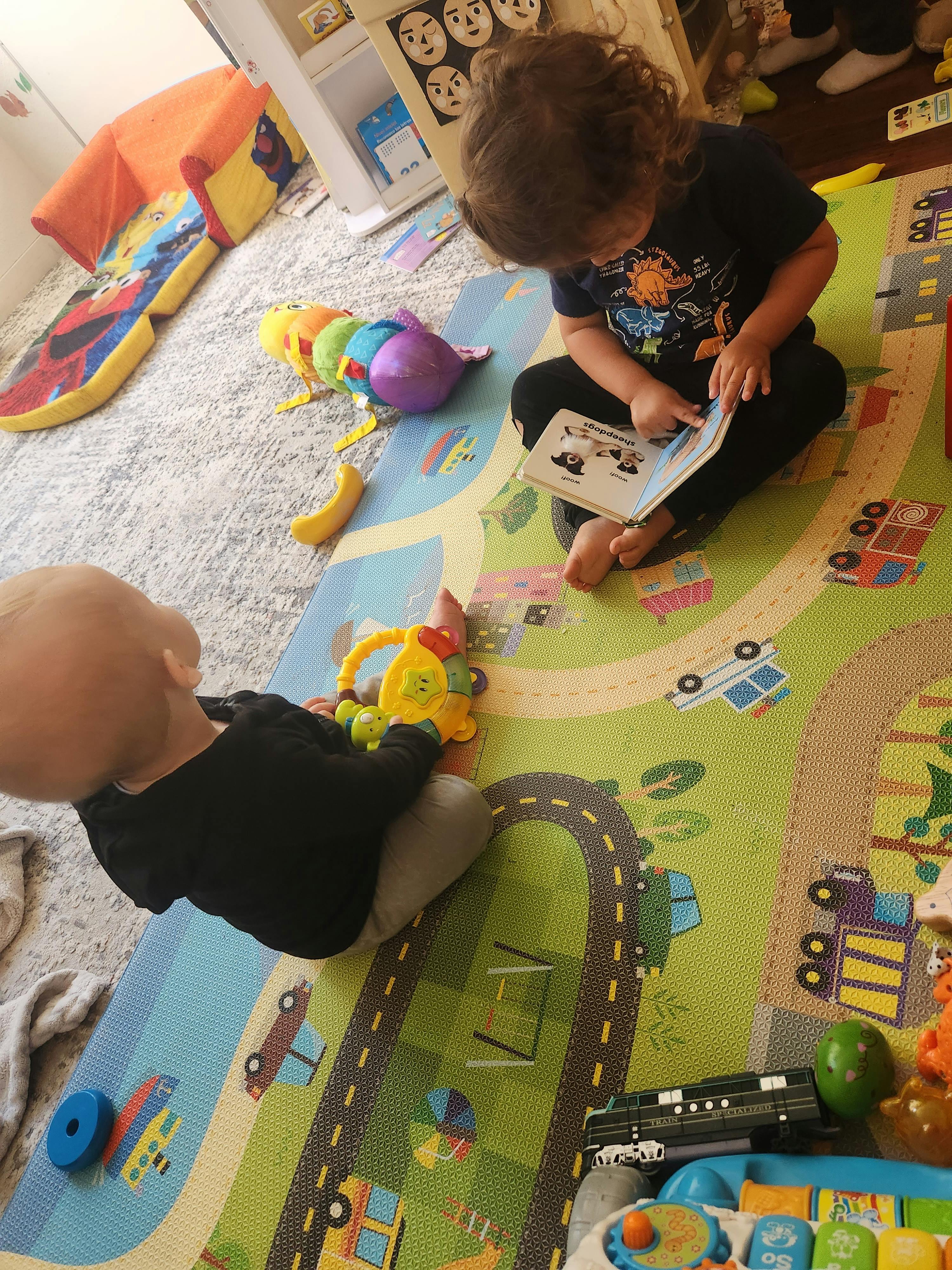 Two children playing on a mat with toys, one reading a book
