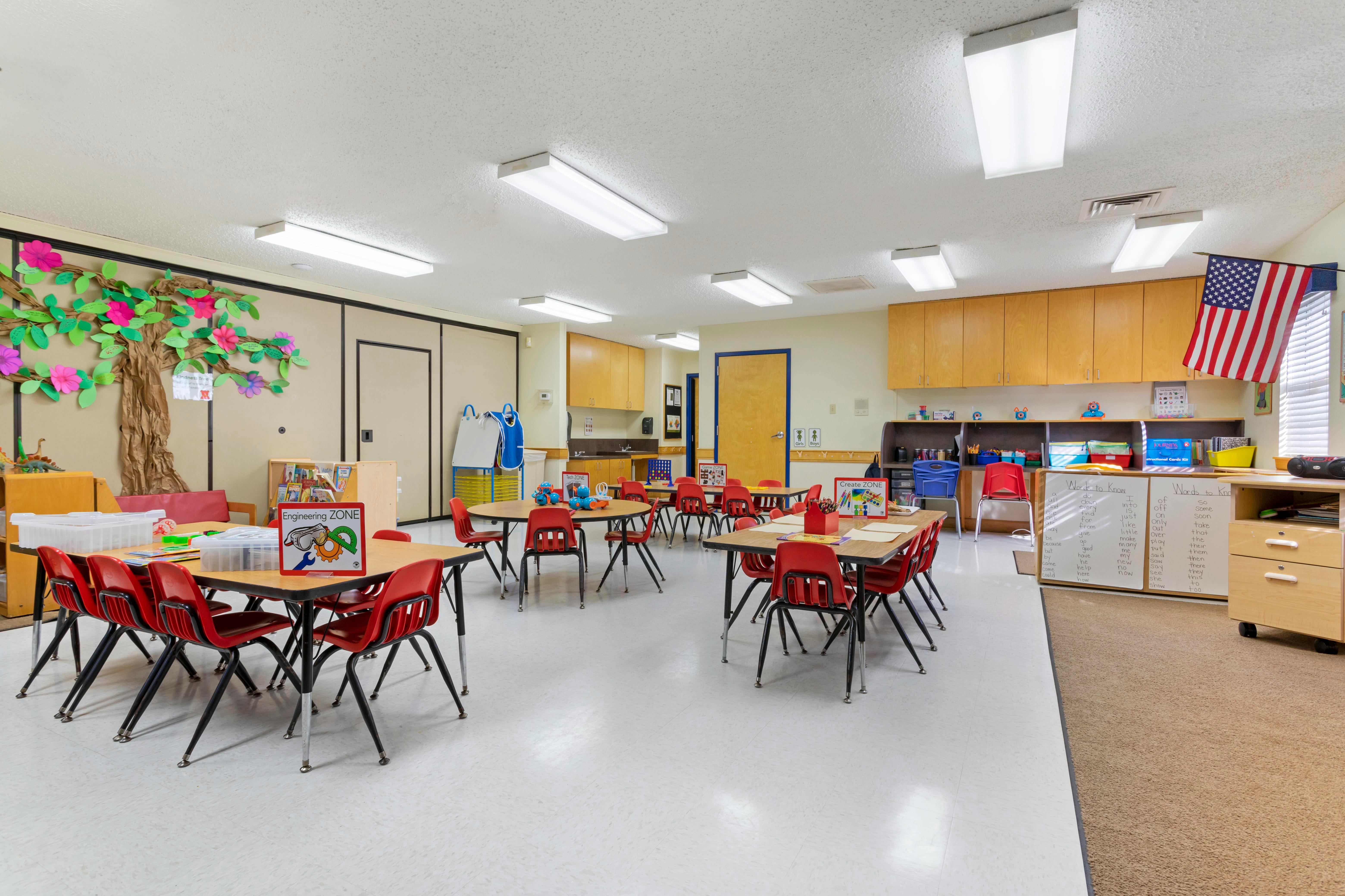An empty classroom with multiple tables and red chairs, an American flag hangs