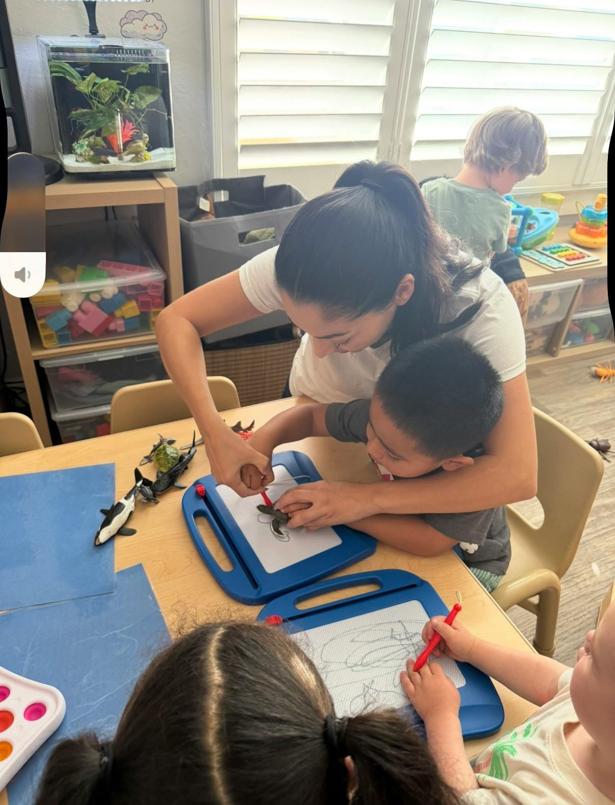Adult helping a child draw on a magnetic board with other children at a table