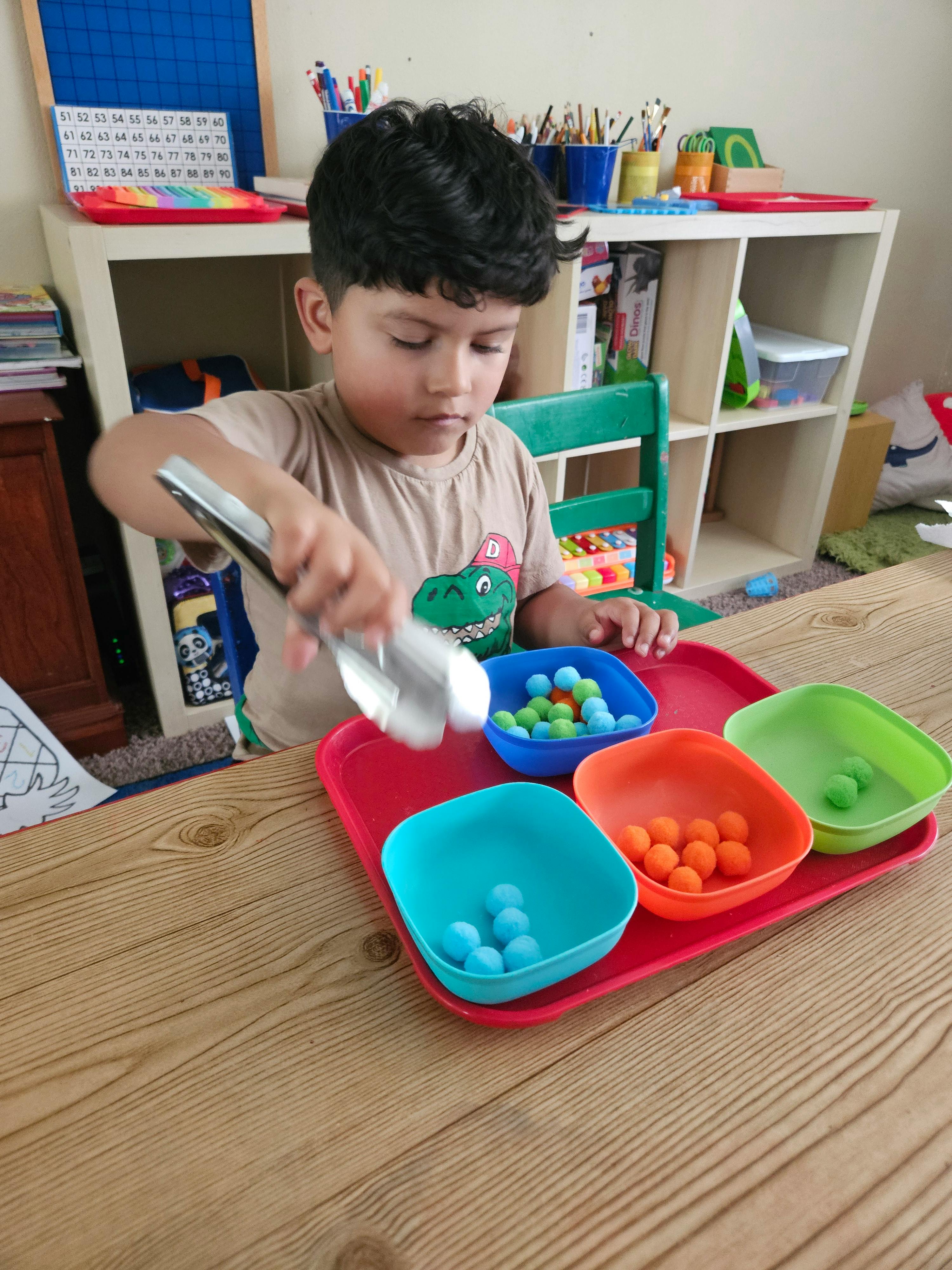 Child using tongs to sort colorful pom-poms into plastic bowls