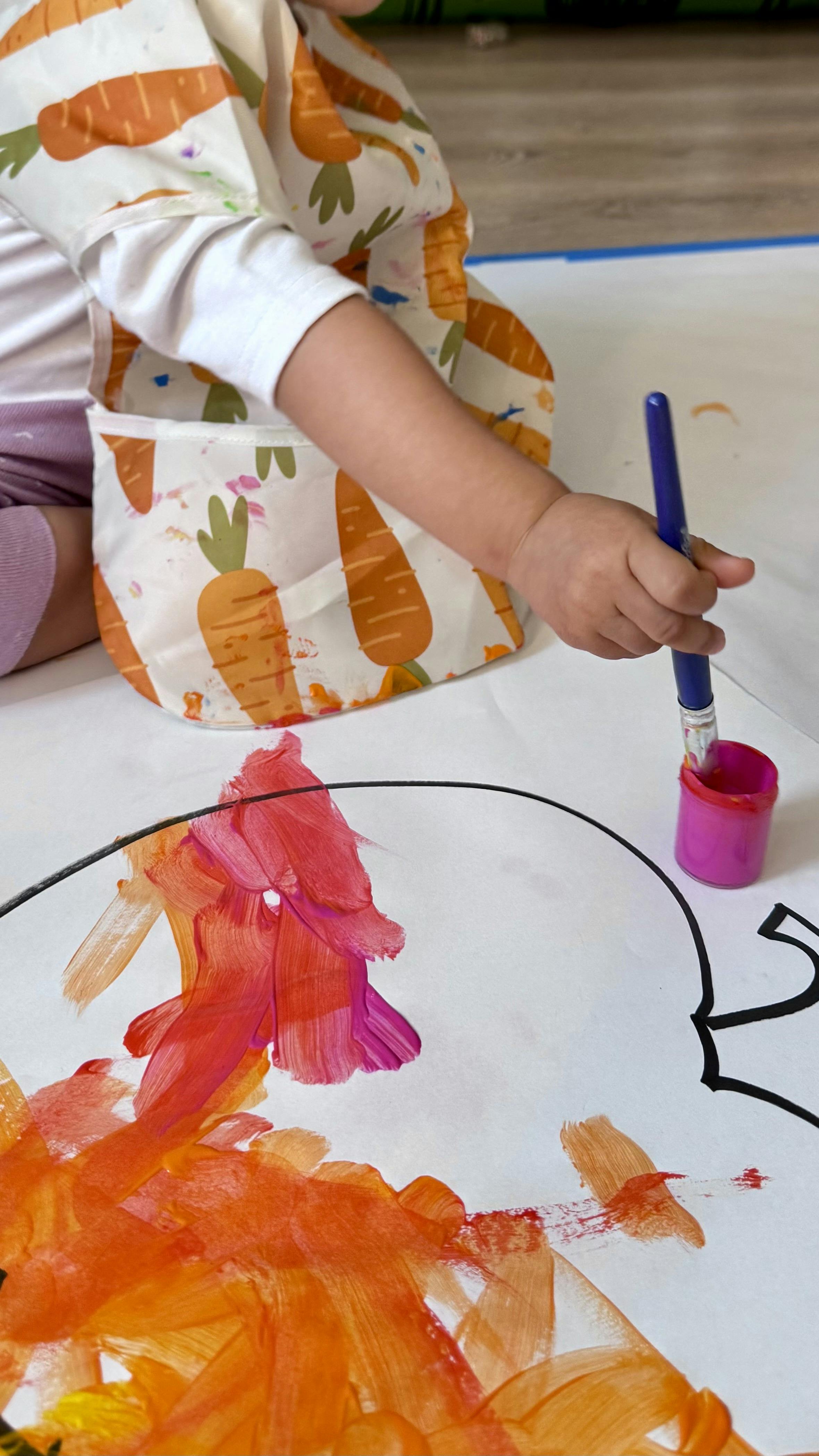 Child in a carrot-patterned bib painting on a large sheet of paper