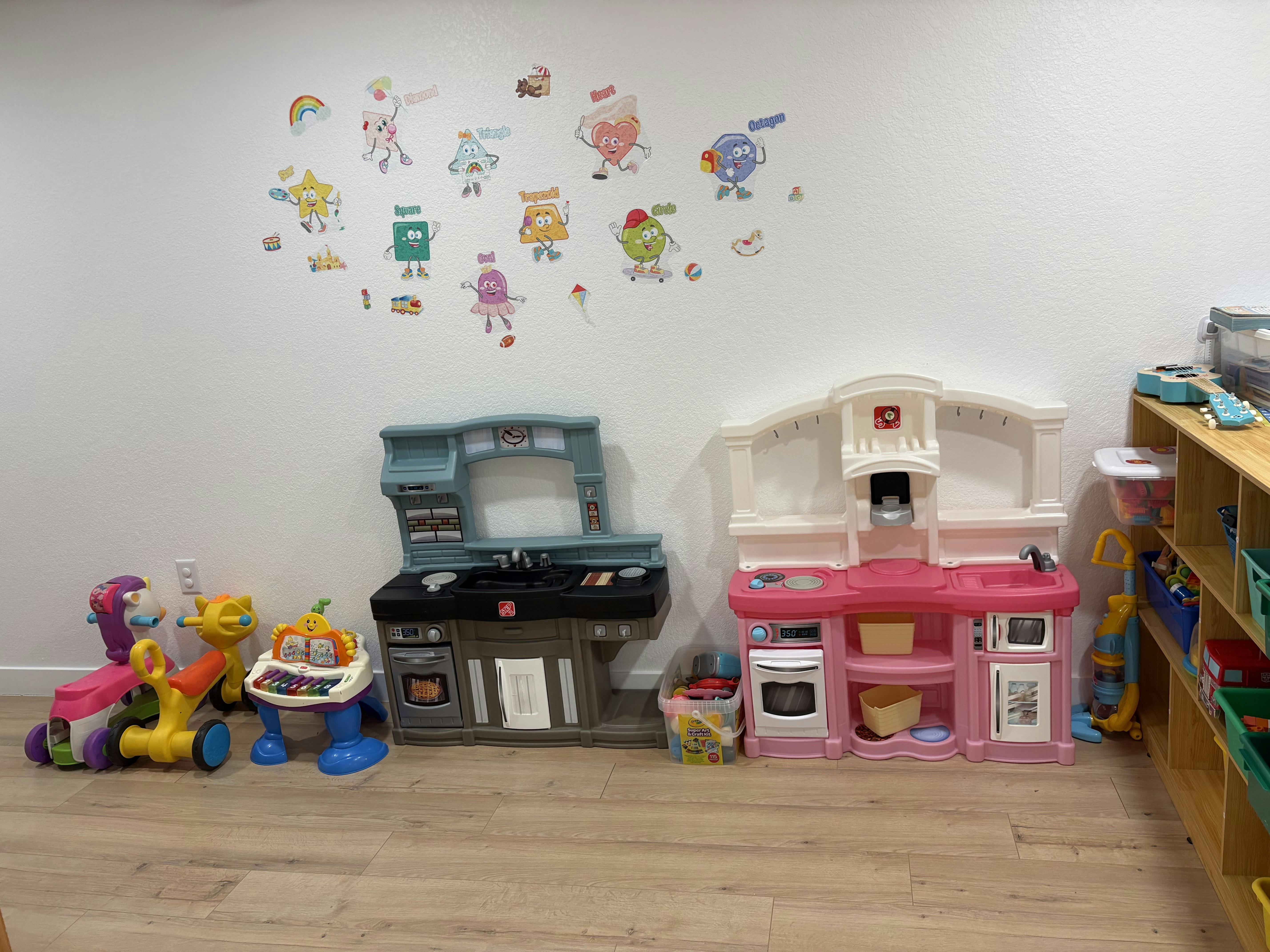 Playroom with two toy kitchens and ride-on toys against a wall with shape decals