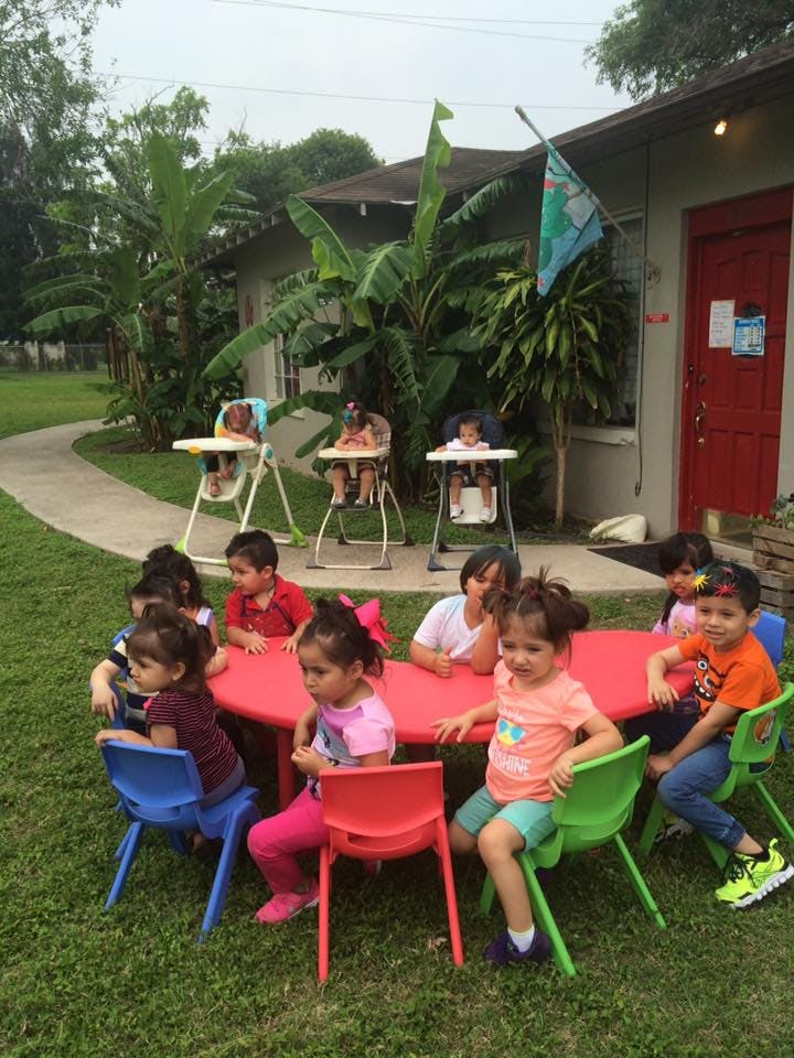 Children sitting at a red table outdoors, some in high chairs, outdoors