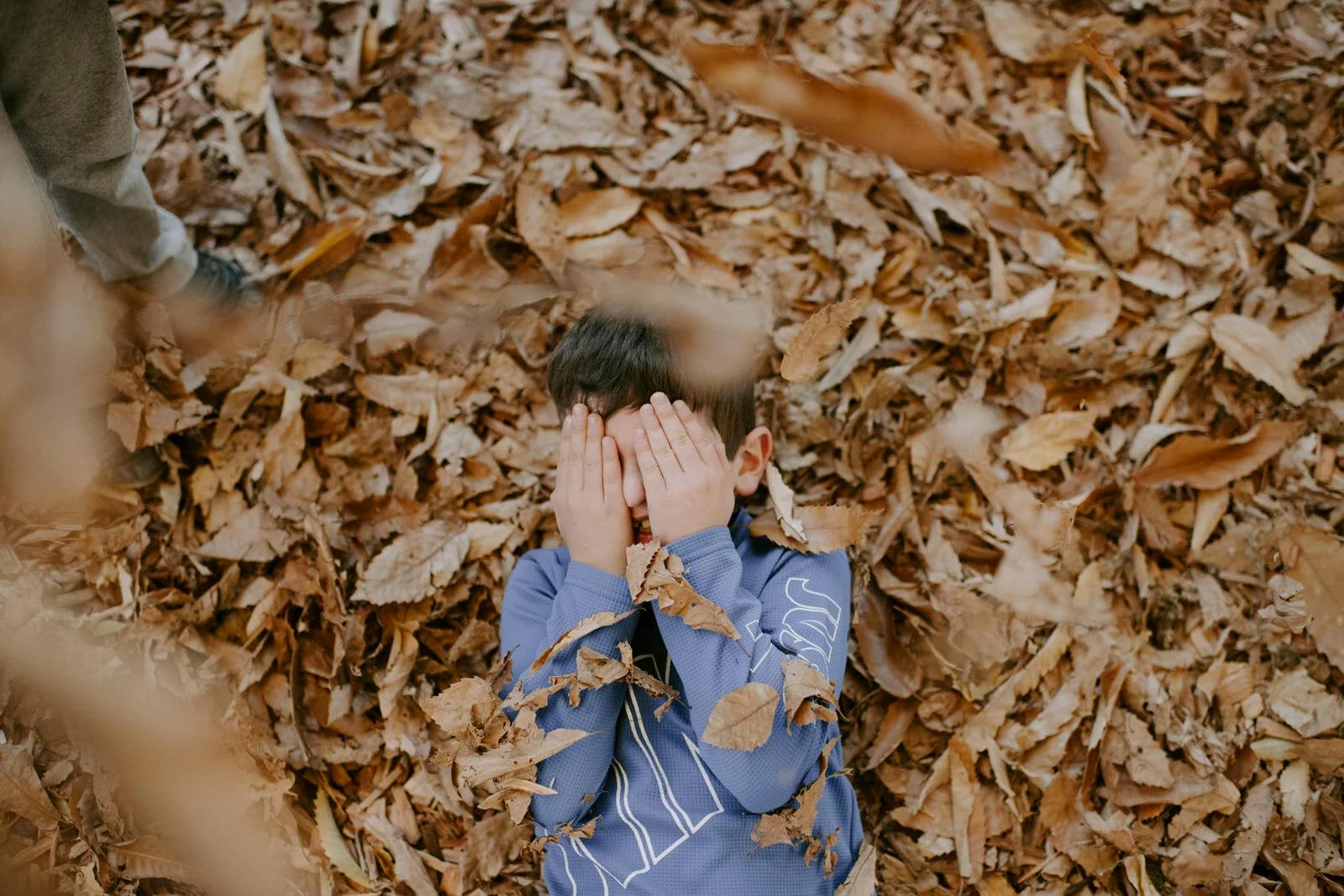 Child lying on a bed of fall leaves covering face with hands