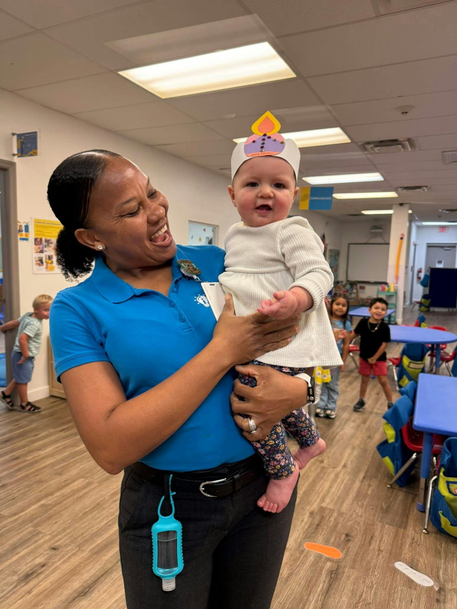 Adult holding a child wearing a paper diya hat in a classroom