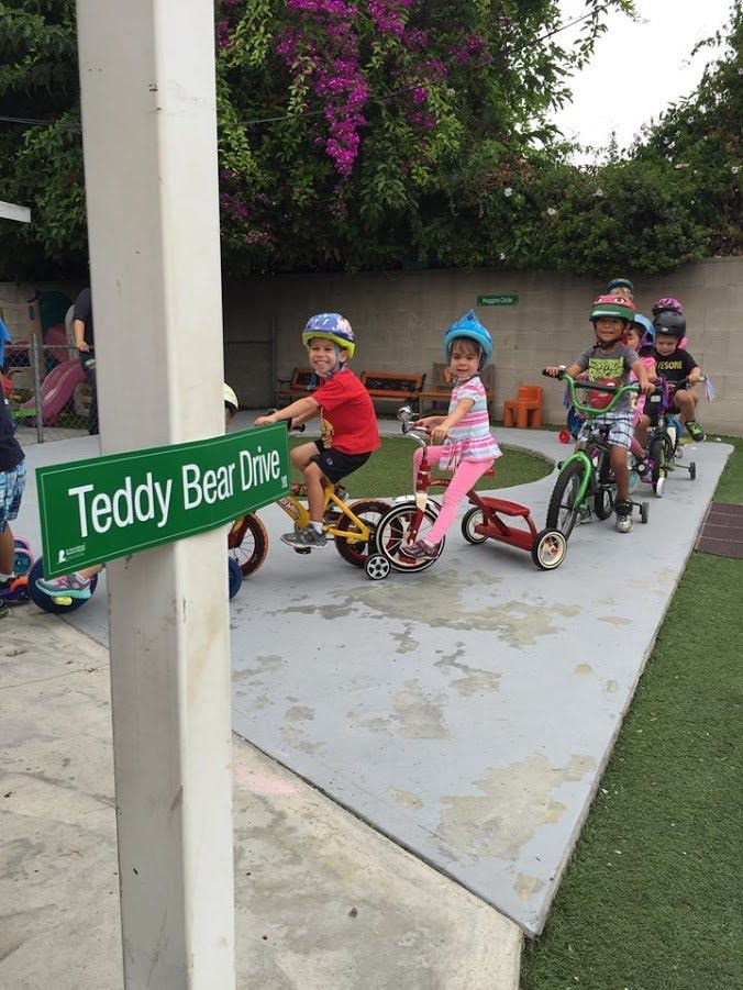 Children on tricycles and bikes with a "Teddy Bear Drive" sign