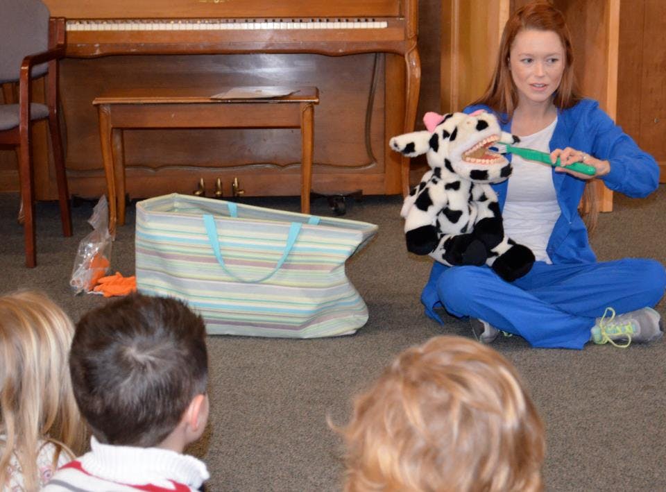 Person demonstrating brushing teeth on a cow puppet to children