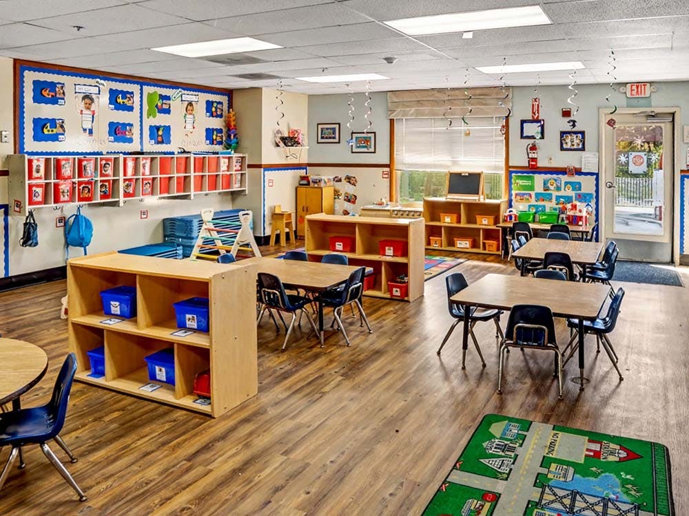 Empty classroom with small tables and chairs, shelves with storage bins, and decorations