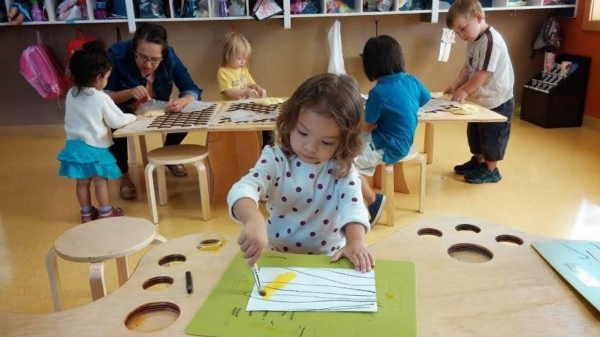Child painting at a table in a classroom with other children and an adult