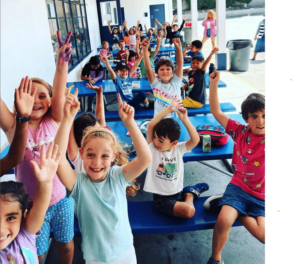 Group of children raising hands while sitting at tables outdoors