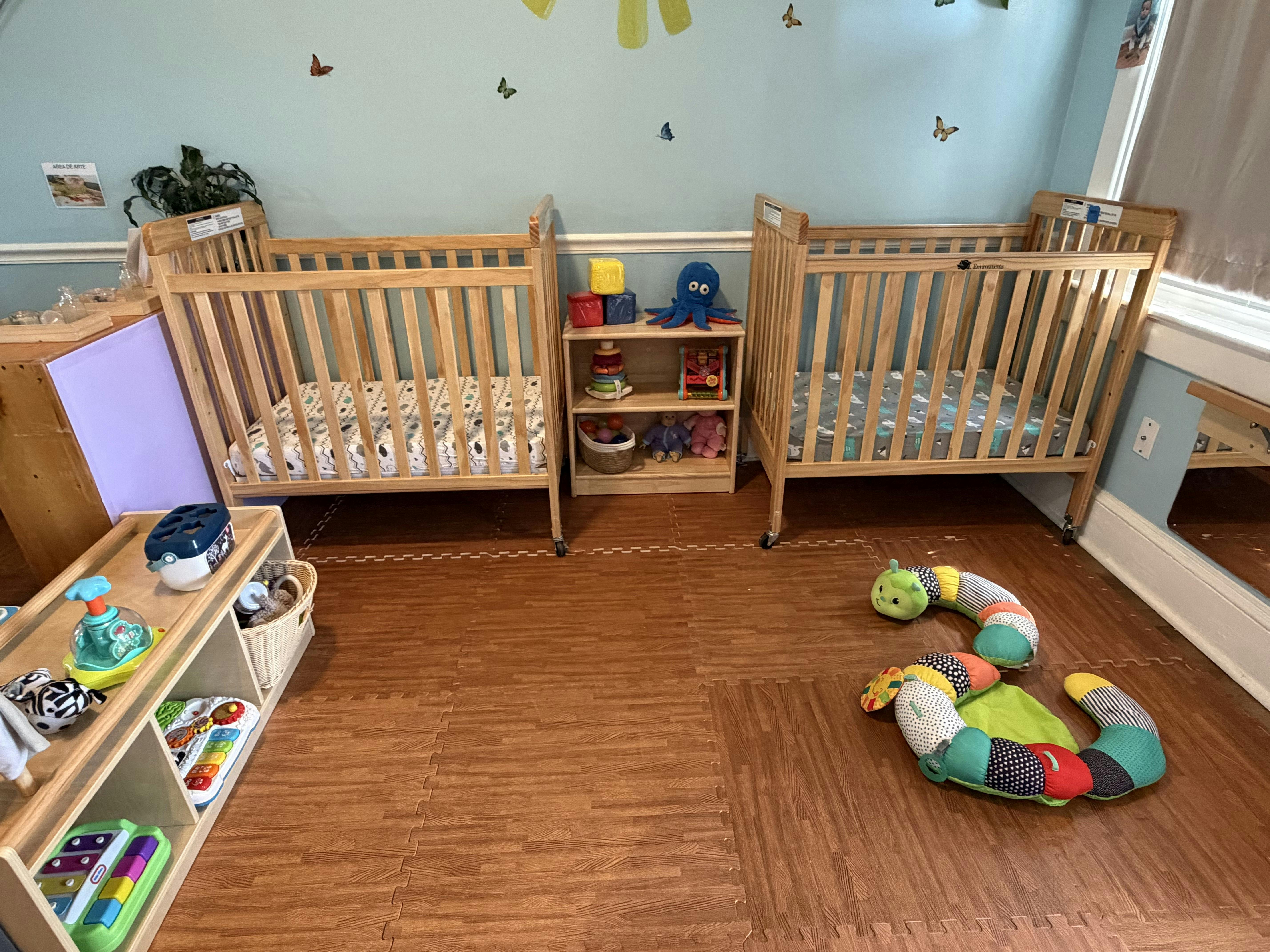 Two wooden cribs separated by a toy shelf in a childcare room