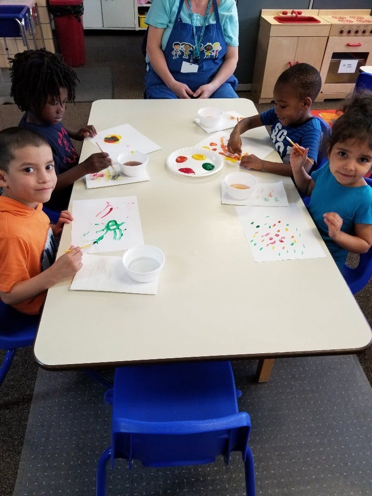 Four children painting at a table with an adult in the background