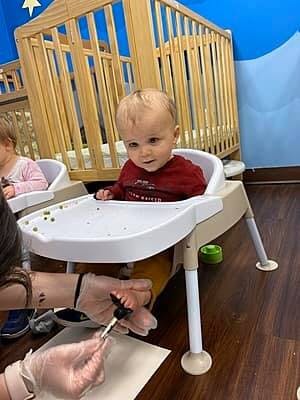 Child in a high chair looking at a person painting their hand