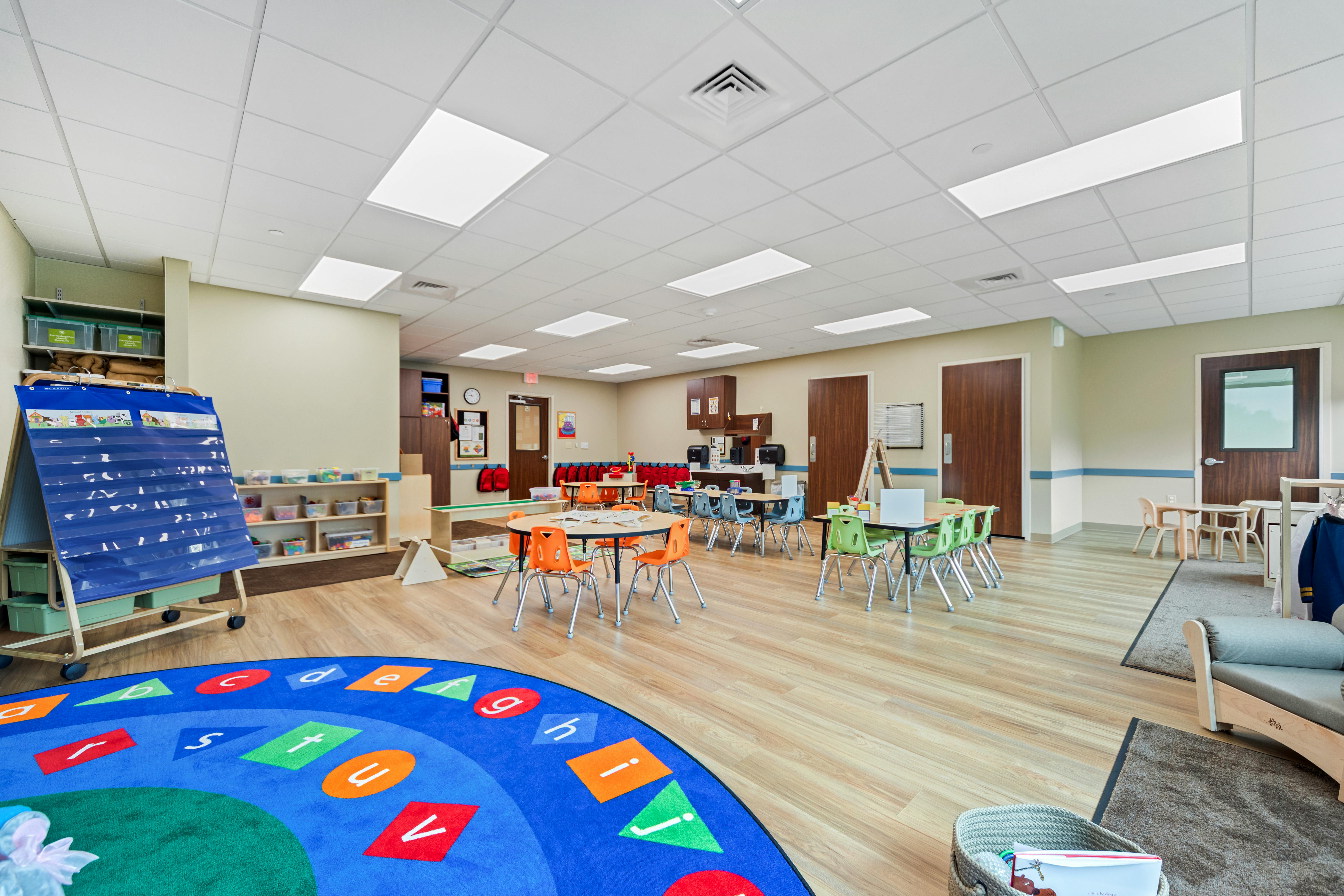 Empty classroom with small tables and chairs, a colorful rug, and storage shelves