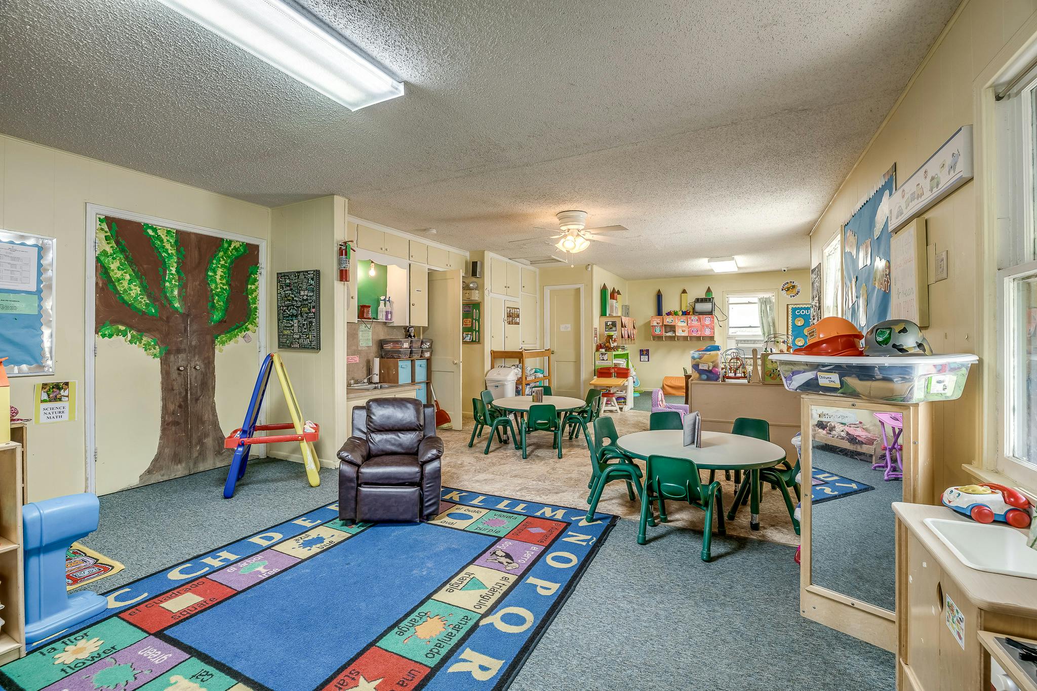 A classroom with small tables and chairs, a large rug, and a tree mural