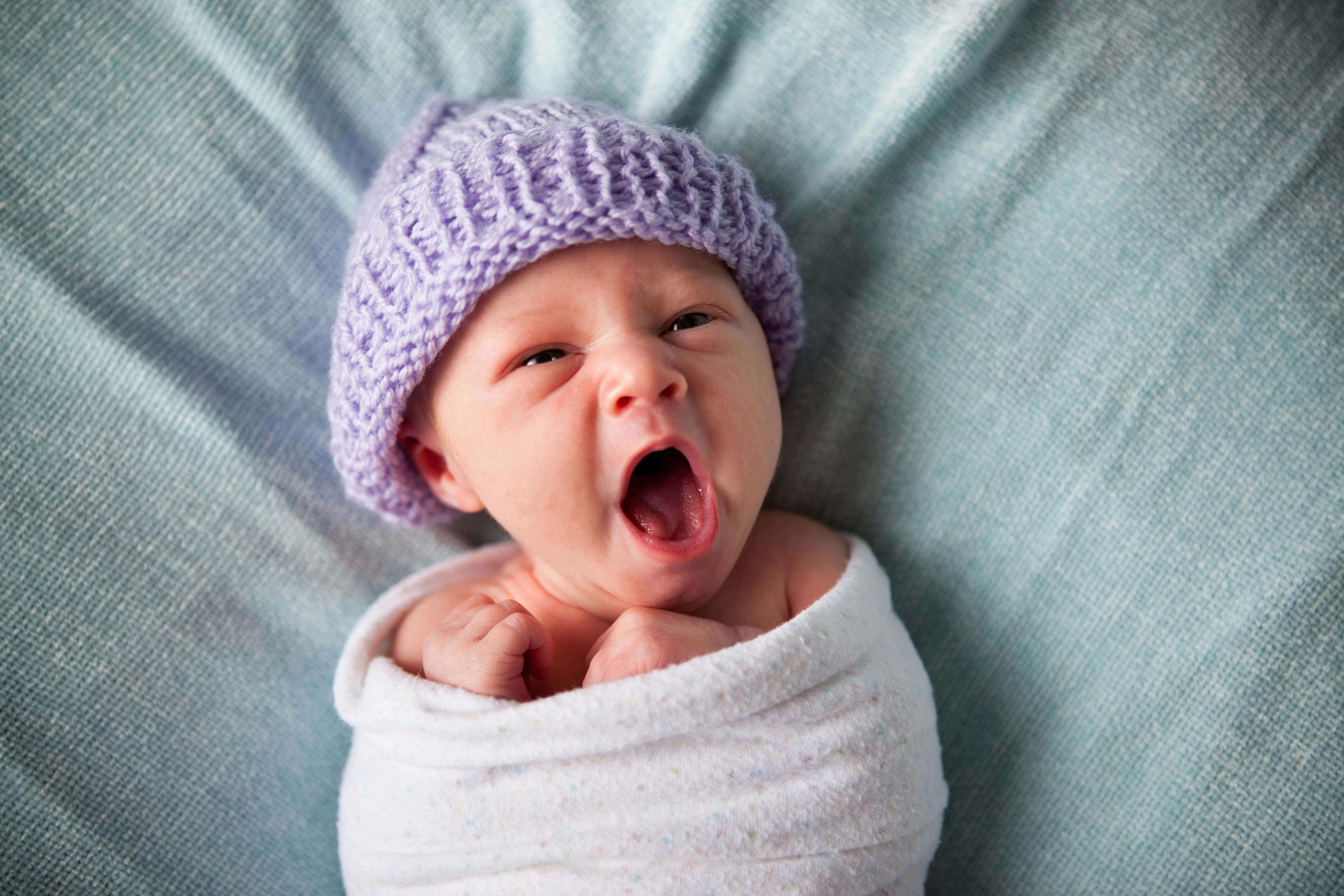 Child in a purple knit hat yawning while swaddled in a white blanket