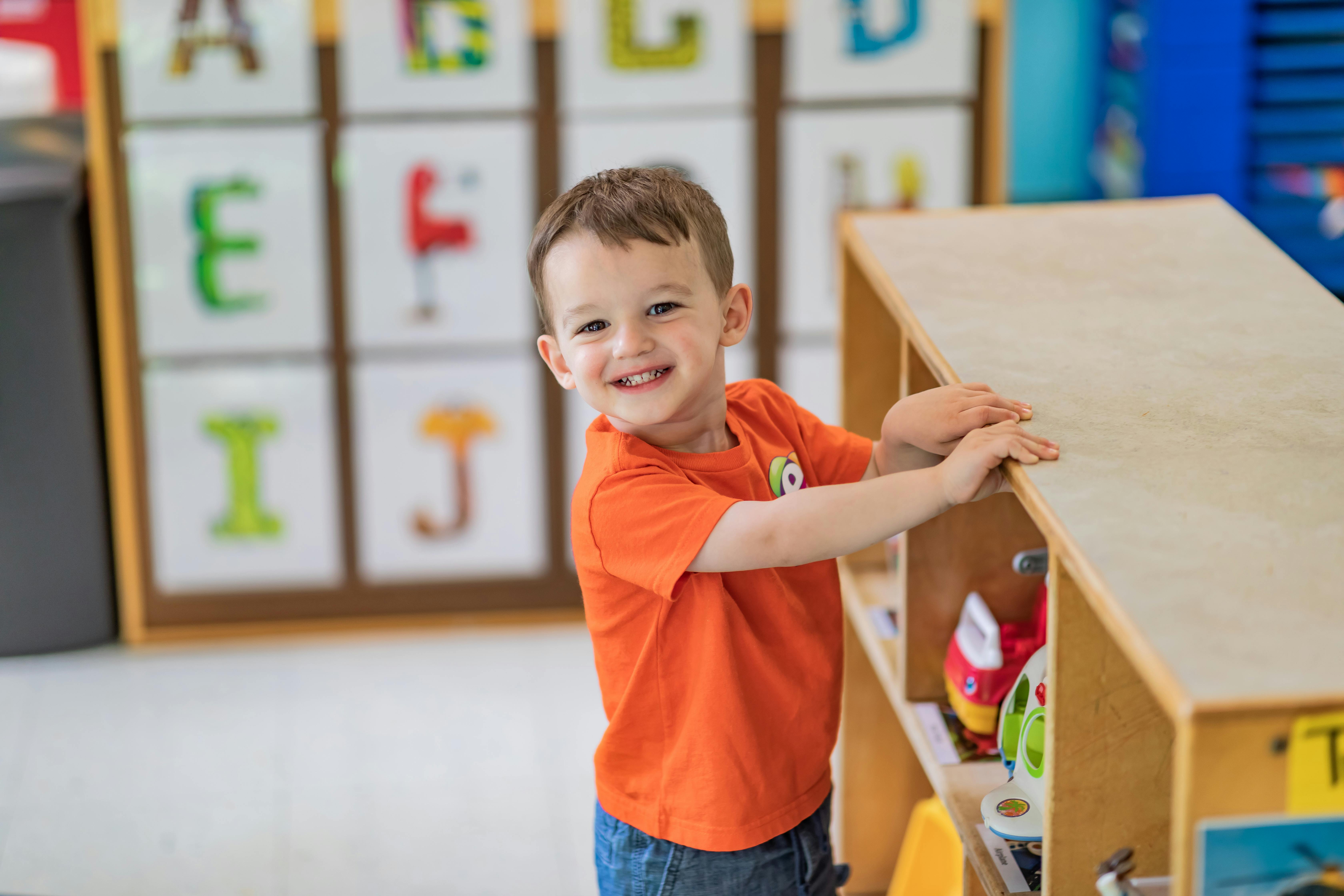 Child in orange shirt standing next to a wooden shelf in a classroom