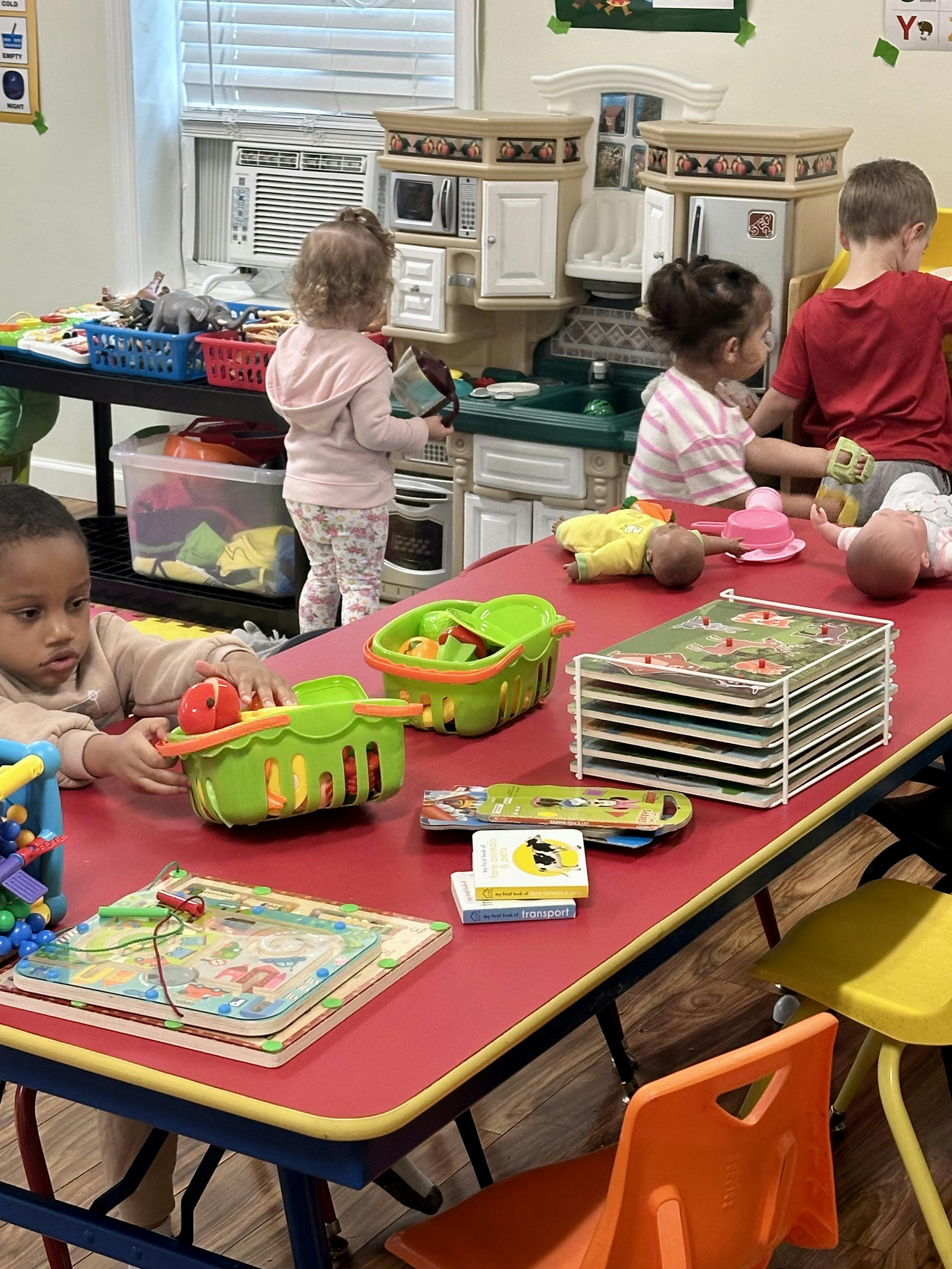Children playing with toy kitchen, dolls, baskets, and puzzles in a classroom