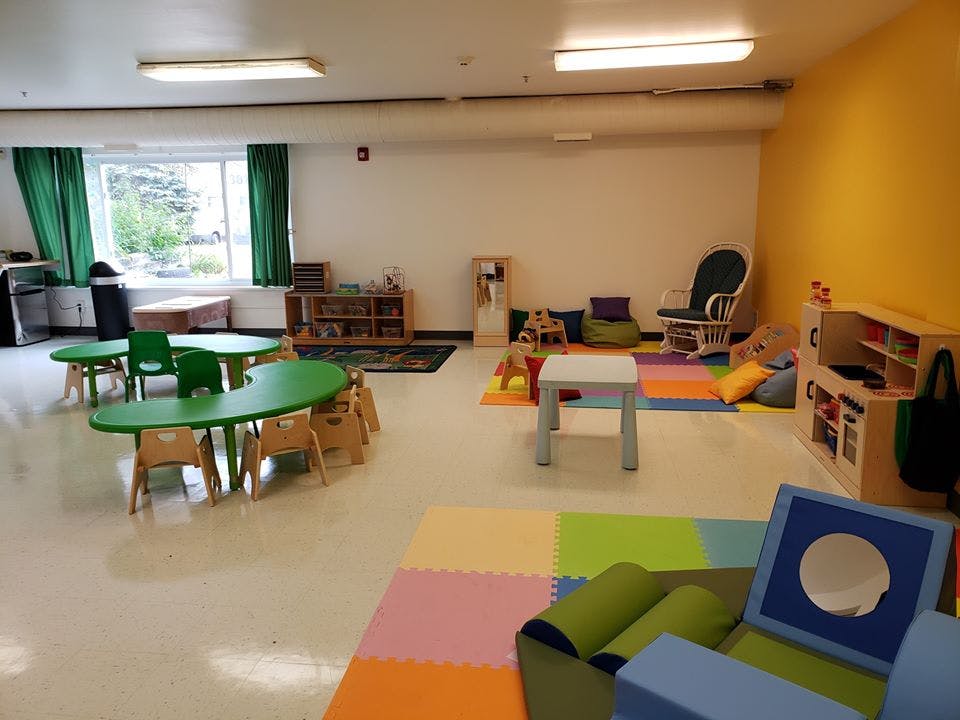 Empty classroom with small green tables and chairs, colorful playmats, and toys