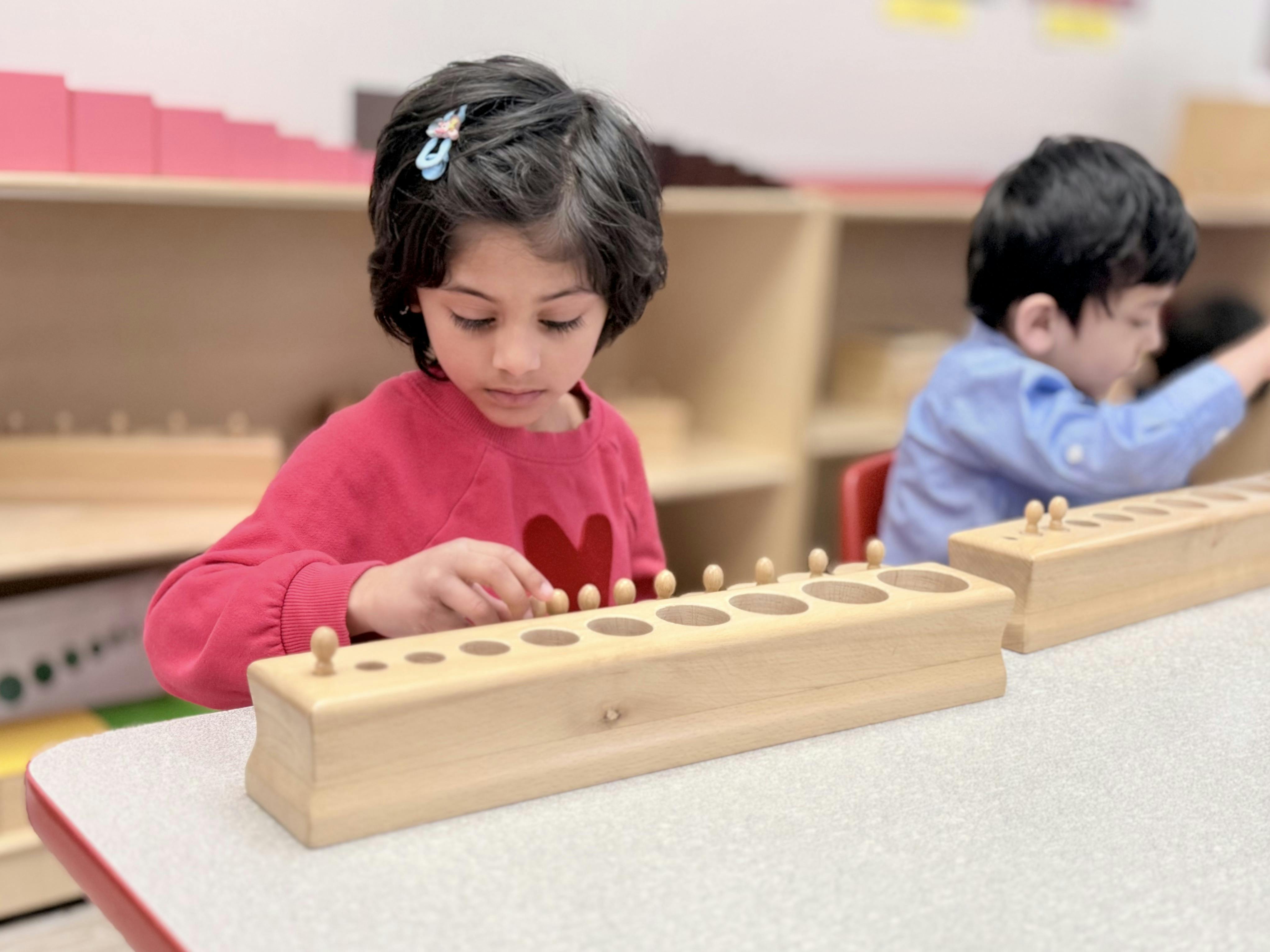 Child in a red sweater placing wooden pegs into a wooden board in a classroom