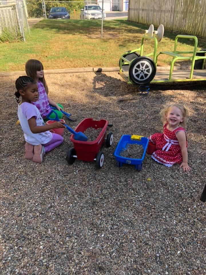 Three children playing with toy wagons and shovels on a gravel surface