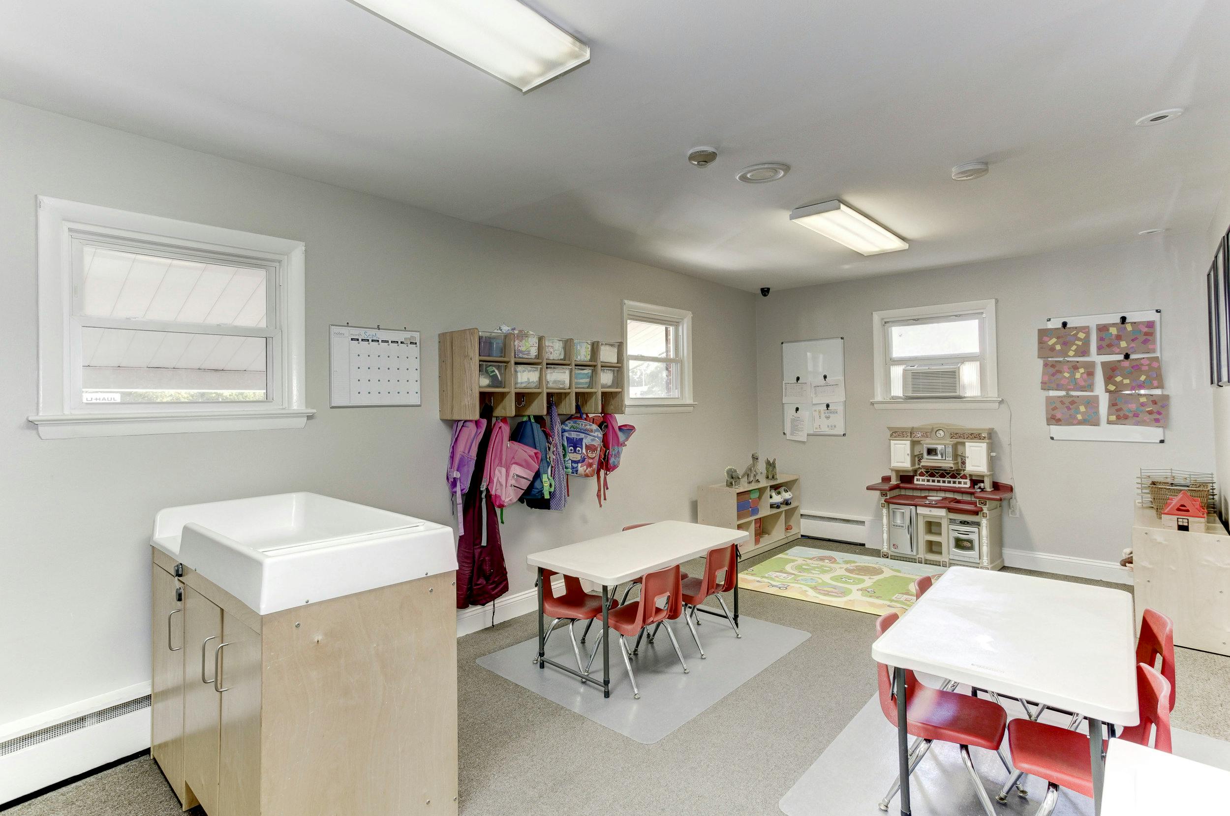 Classroom with small tables and chairs, cubbies with backpacks, and a changing station