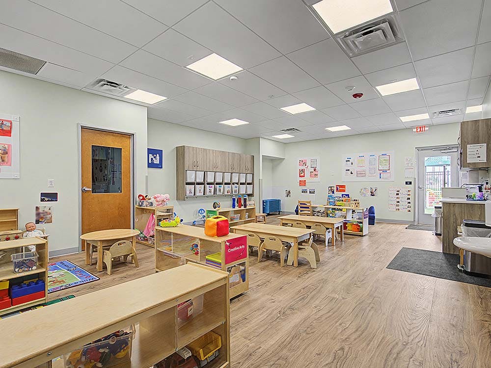 An empty childcare classroom with small tables, chairs, and shelves of toys