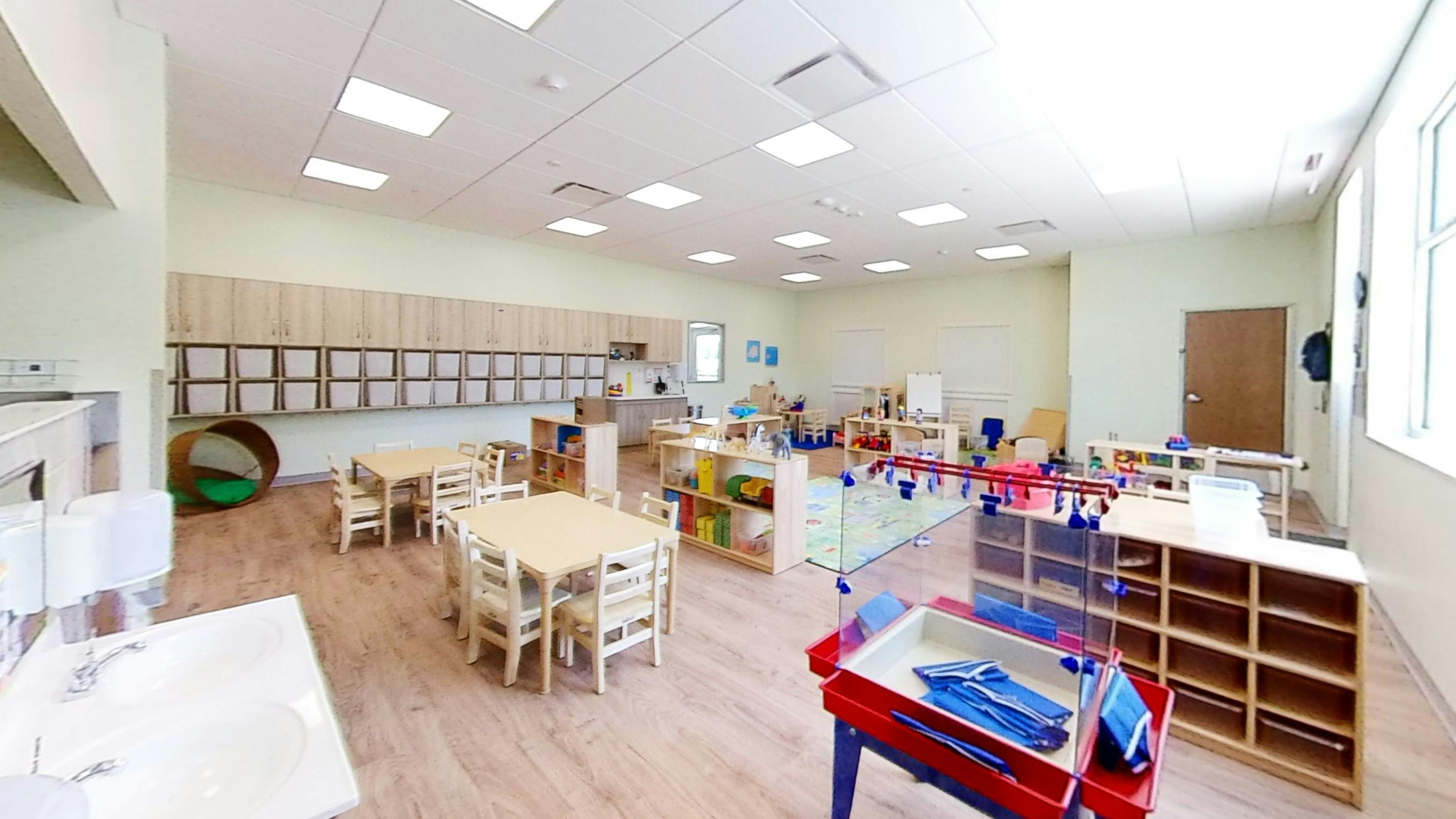 An empty bright classroom with small tables, chairs, shelving units, and a sink