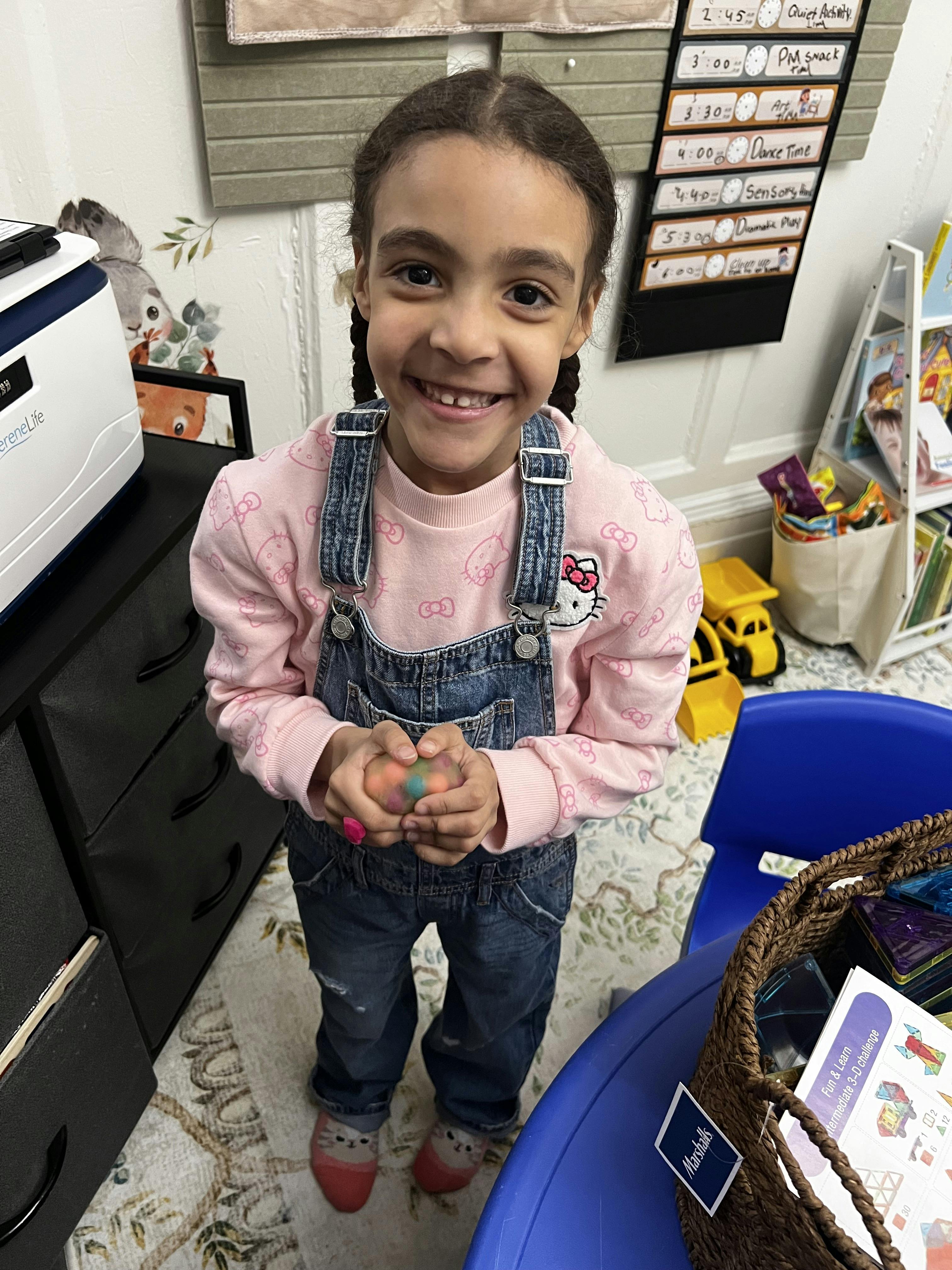 Child in denim overalls and pink shirt holding a colorful stress ball