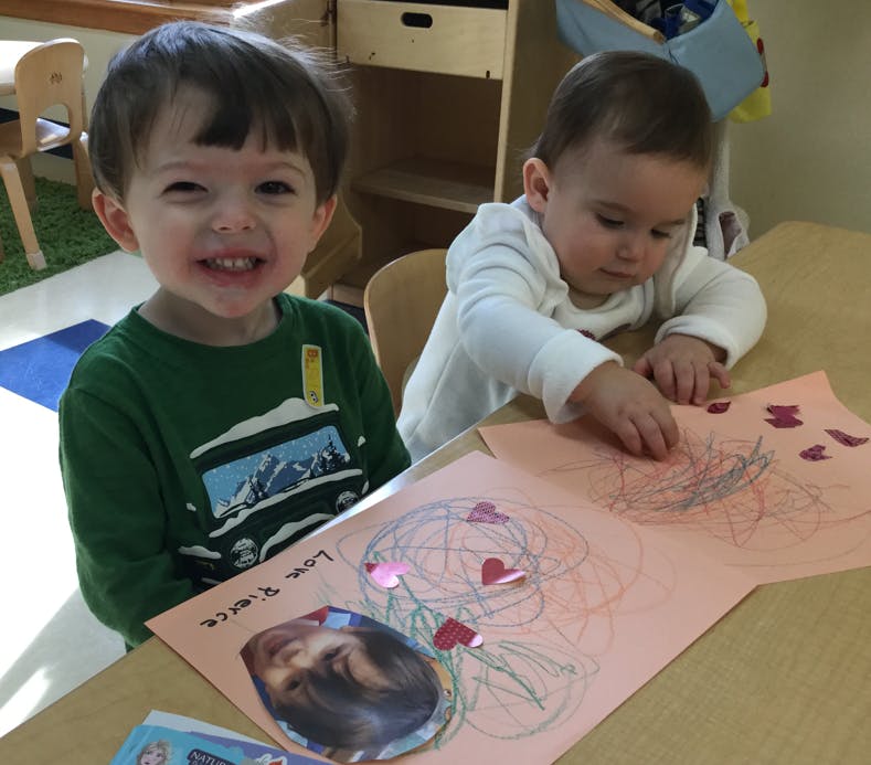 Two children at a table, one smiling at the camera, the other drawing