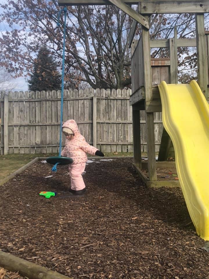 Child in a snowsuit standing next to a swing in a playground
