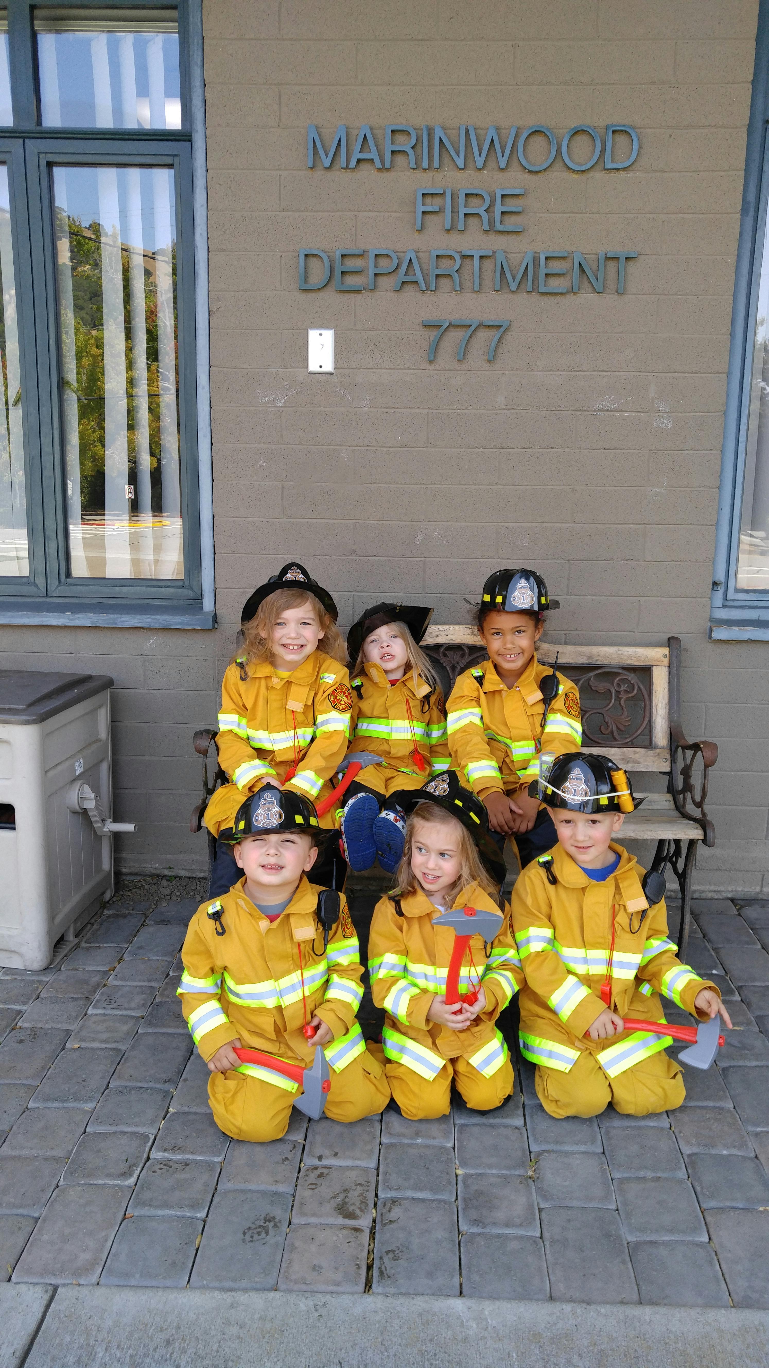 Children in firefighter costumes with toy axes outside a fire department