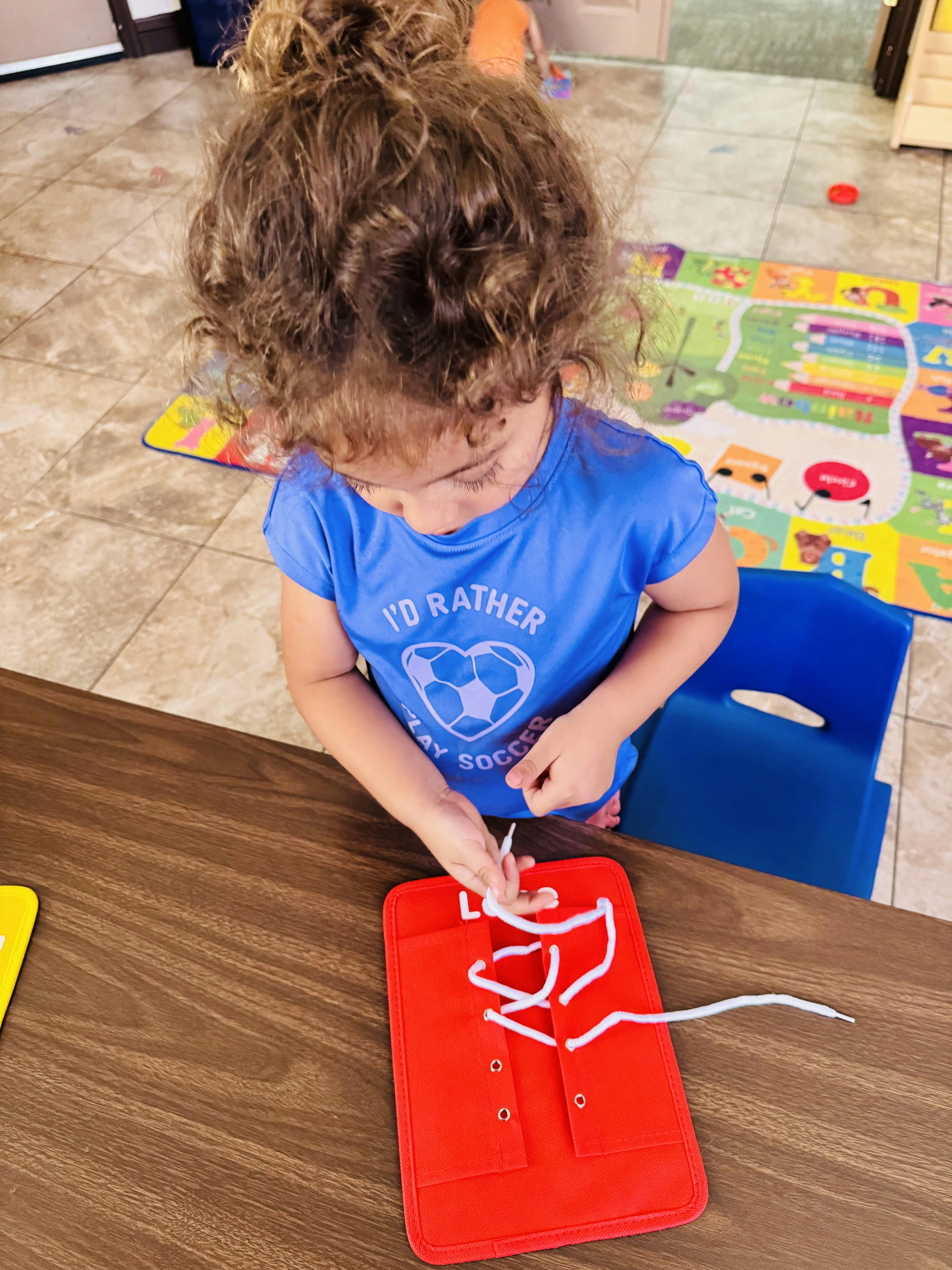 Child in a blue shirt with "I'D RATHER PLAY SOCCER" on it, lacing a red toy