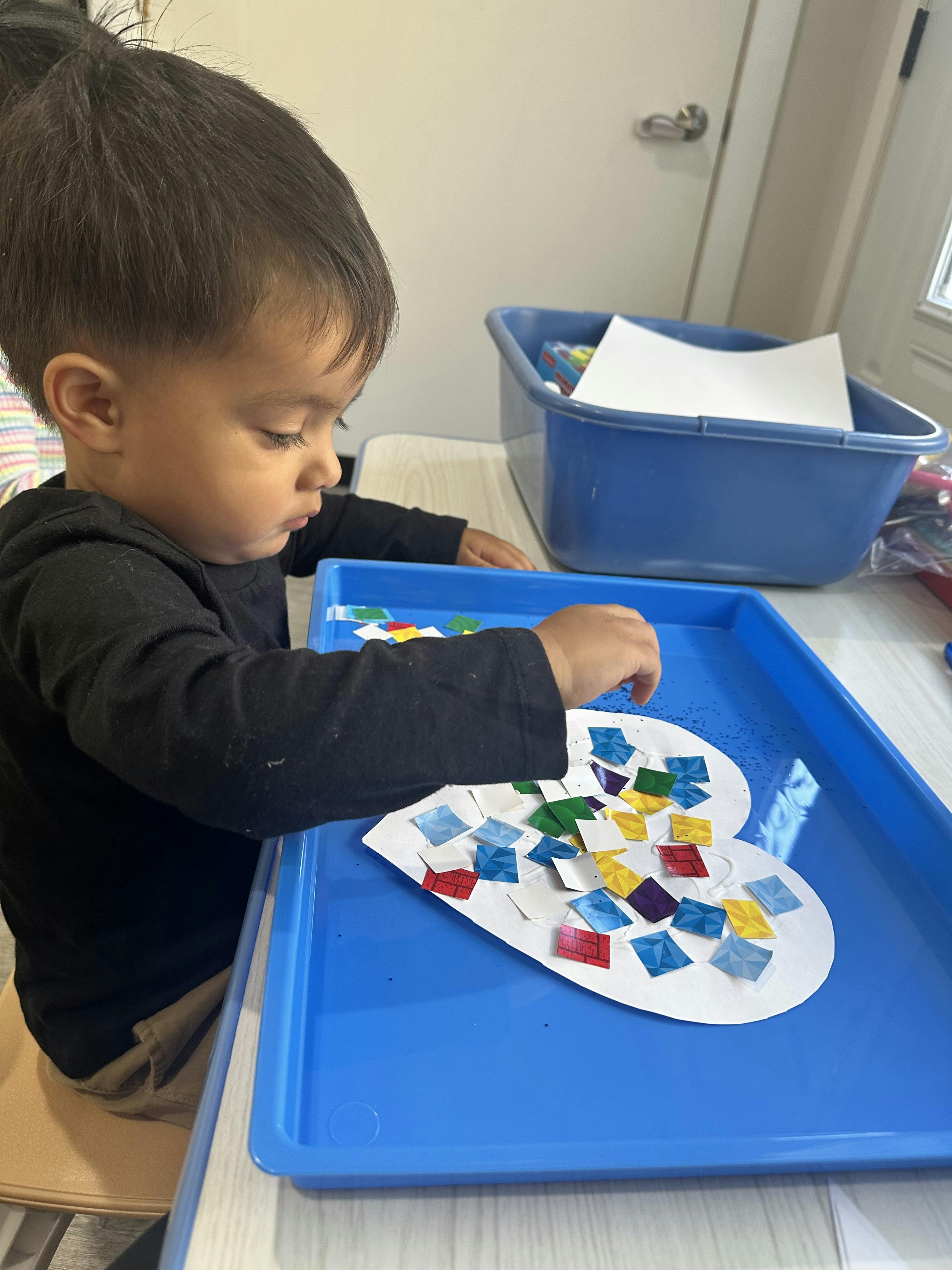 Child gluing colorful paper squares onto a heart-shaped cutout