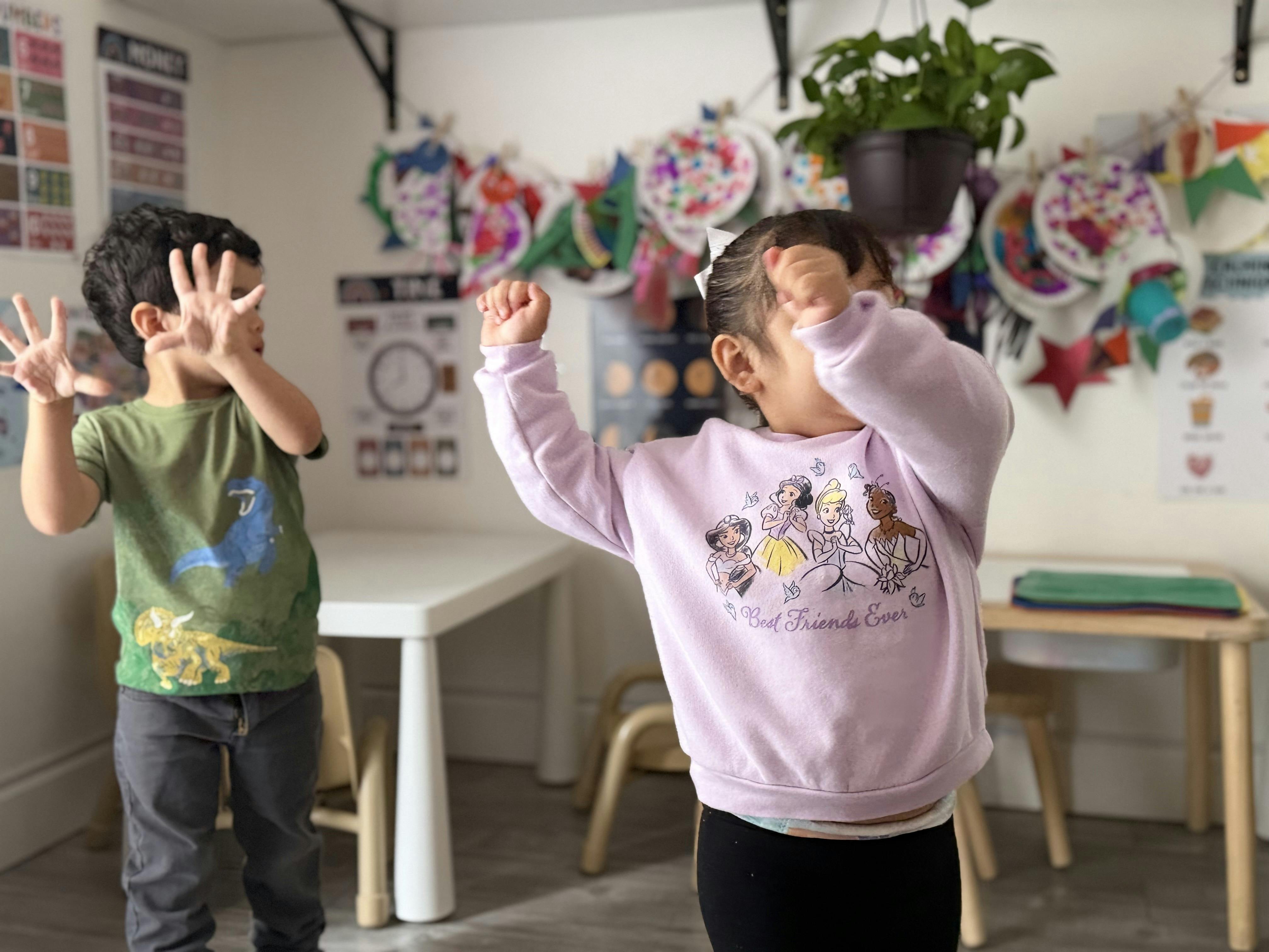 Two children dancing and gesturing in a playroom with crafts on the wall
