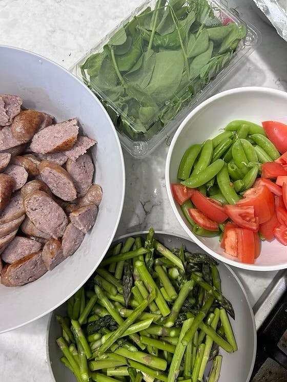 Bowls of sliced sausage, asparagus, tomatoes, and snap peas with a container of spinach