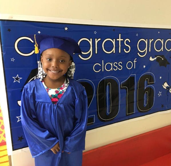 Child in a blue graduation cap and gown in front of a "Class of 2016" banner