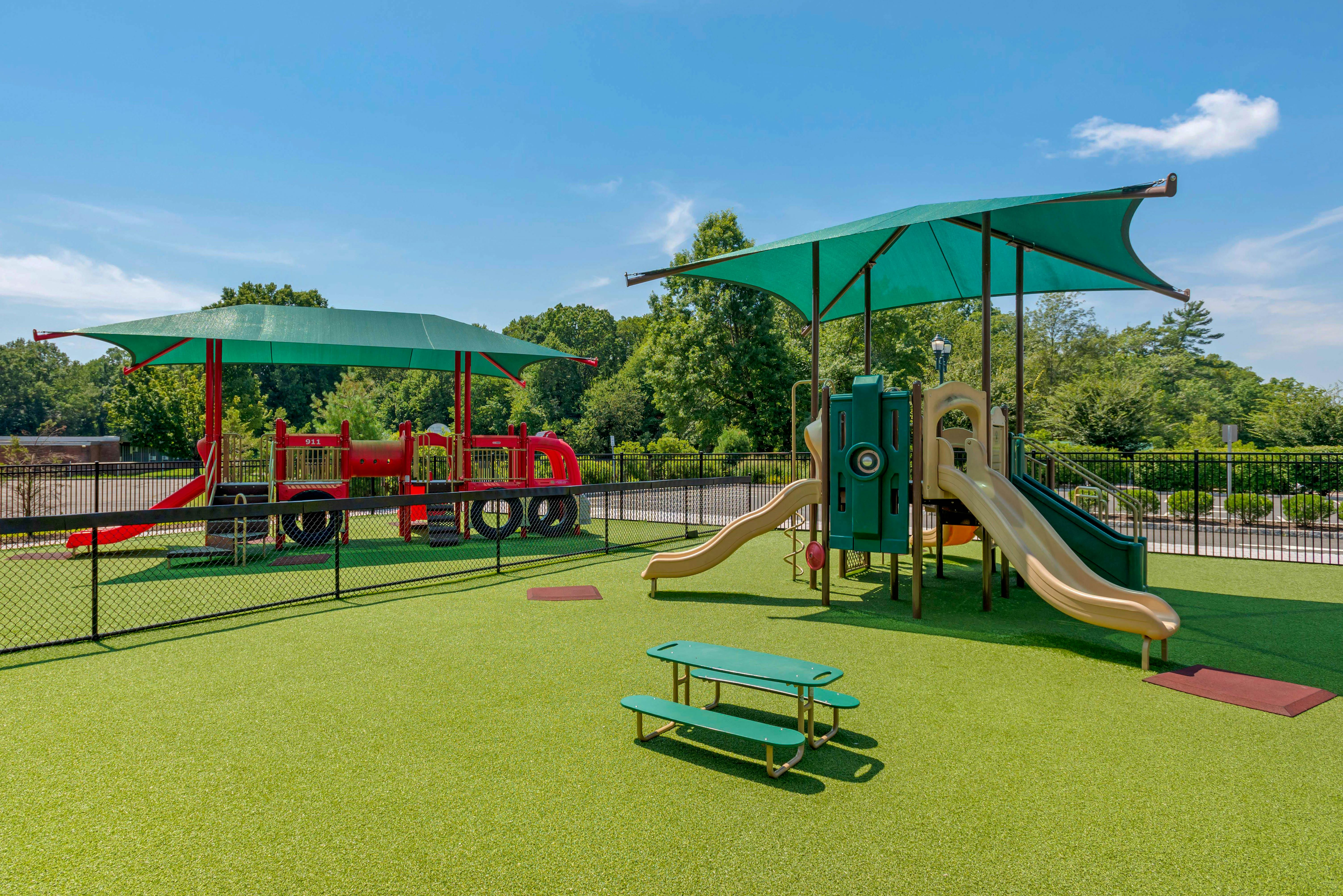 A fenced playground with a red fire truck-shaped play structure and a large play structure with slides and shade