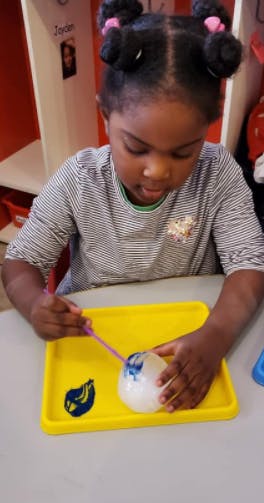Child painting a white object with blue paint on a yellow tray