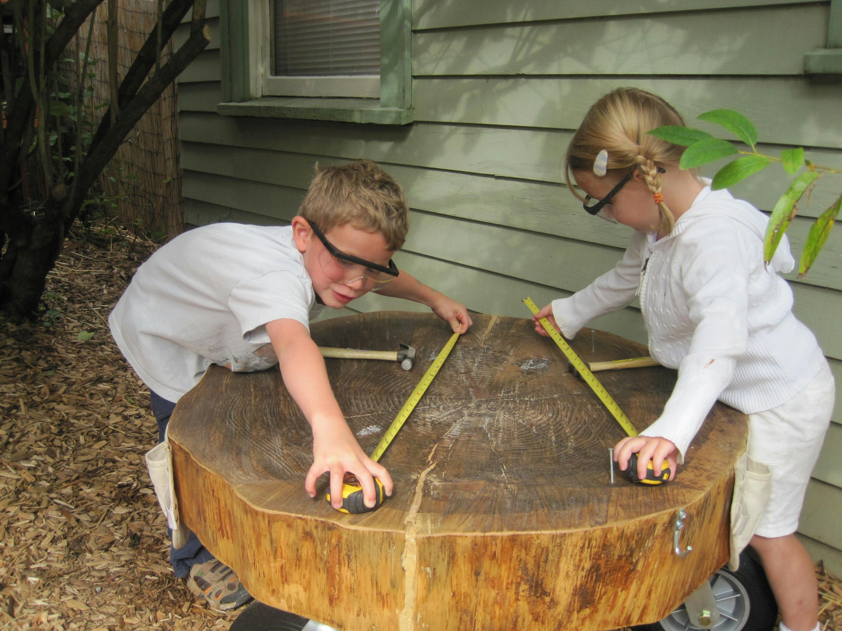 Two children with safety glasses measuring a tree stump with tape measures