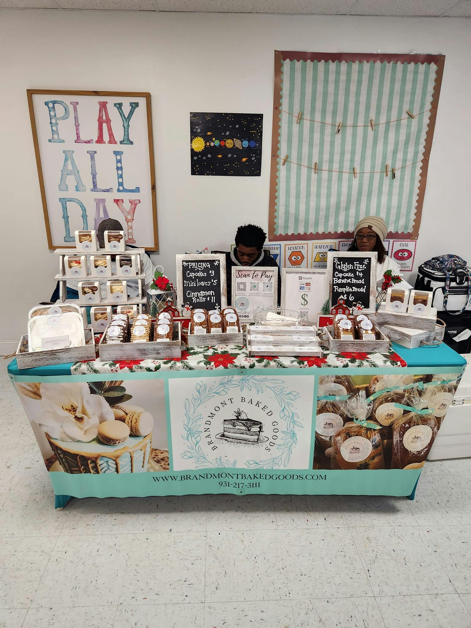People behind a table with baked goods, pricing signs, and a "Scan to Pay" sign