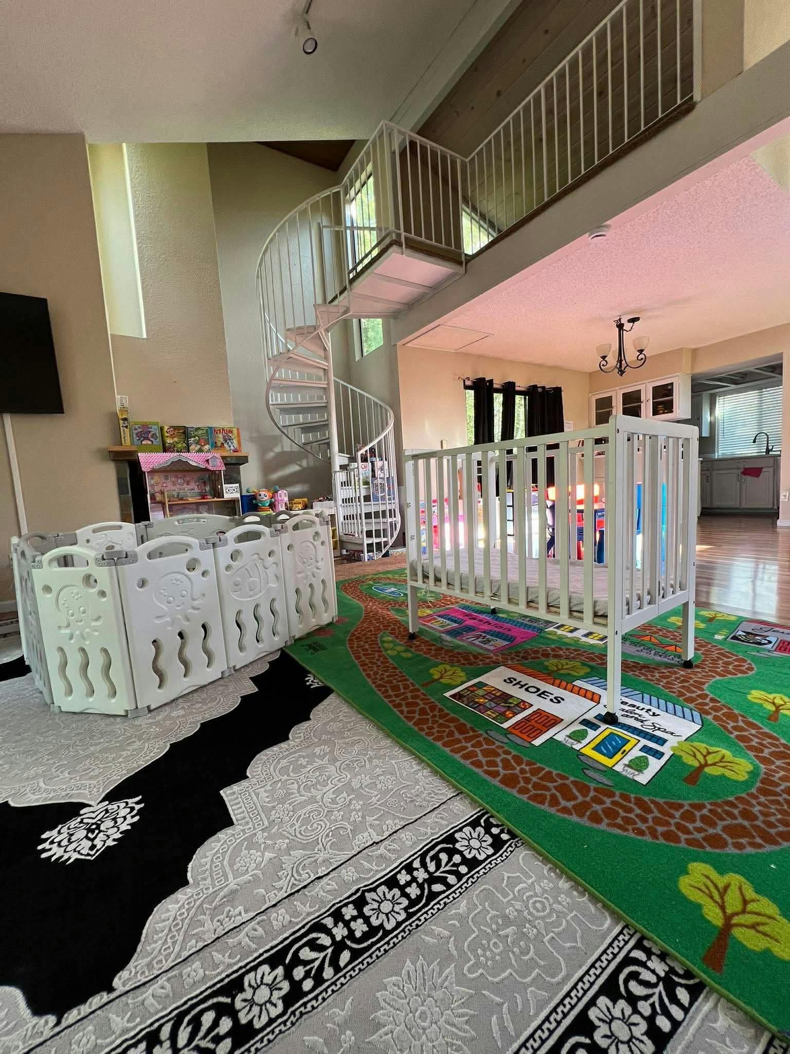 Baby playpen and crib on a colorful rug in a living area with a spiral staircase